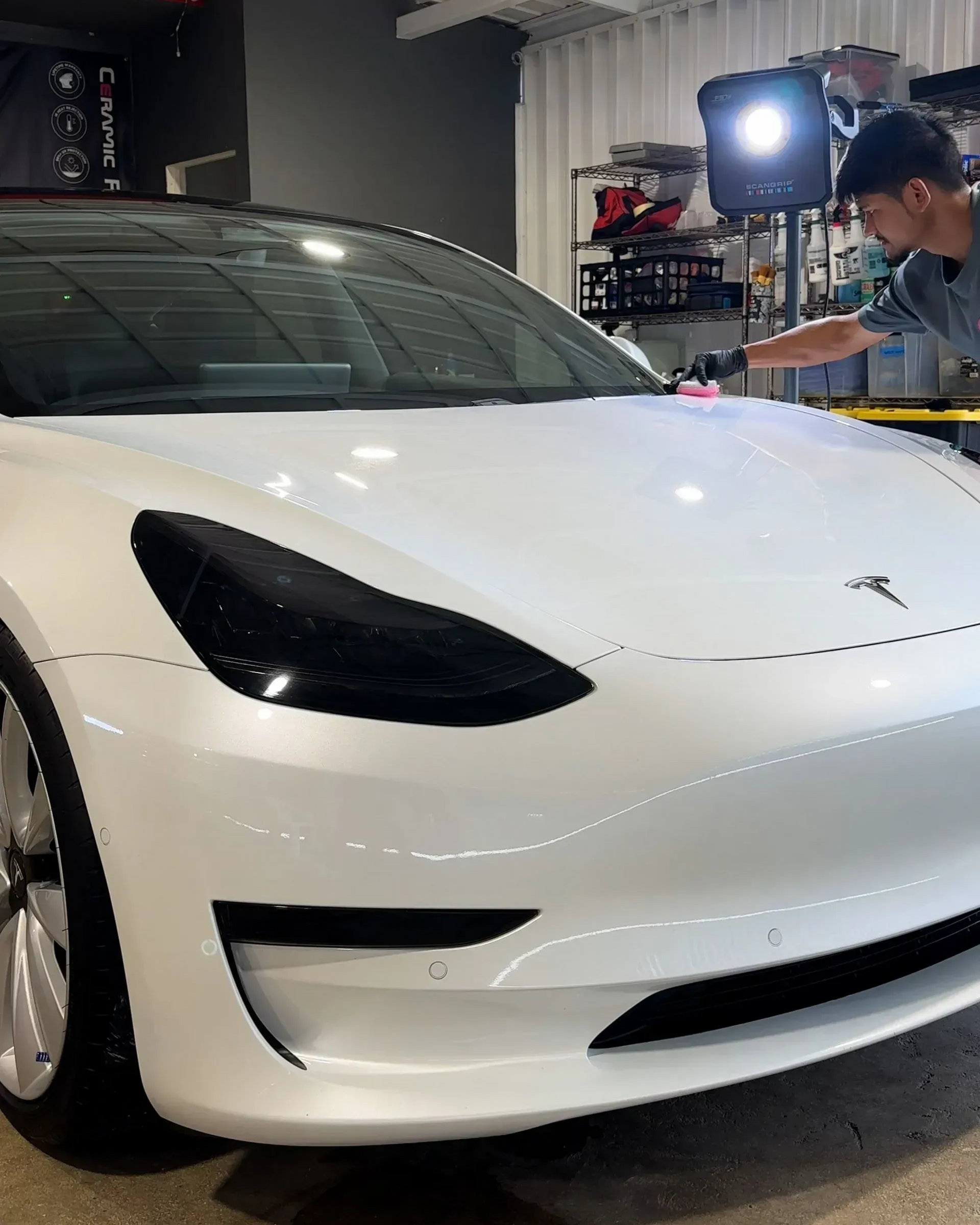 A technician in a garage applies a coating to the hood of a white Tesla with black tinted headlights.