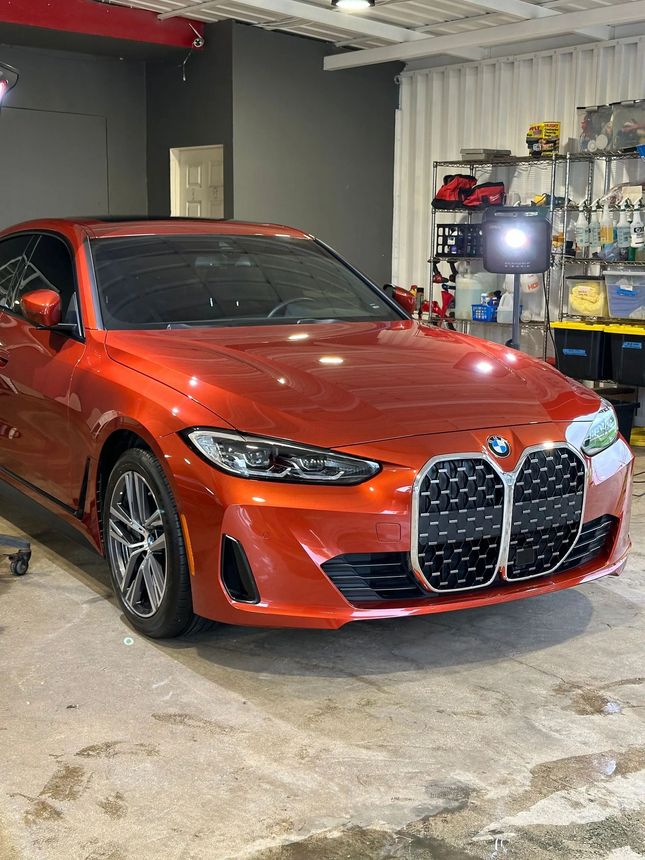 Metallic orange BMW coupe with a large vertical grille parked inside a workshop with shelving in the background.