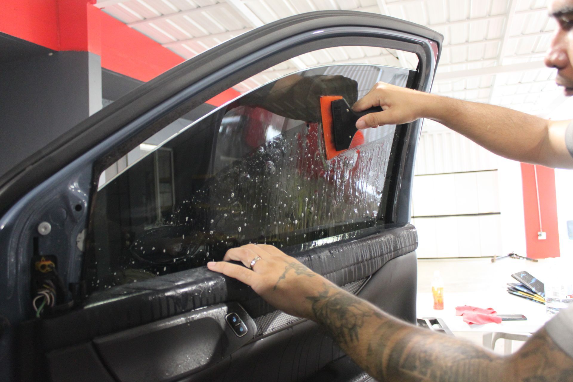 A worker uses a squeegee to apply window tint film to a car door window in a shop.
