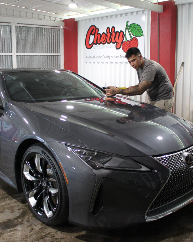 A person buffs the hood of a metallic gray Lexus parked inside a shop with a 
