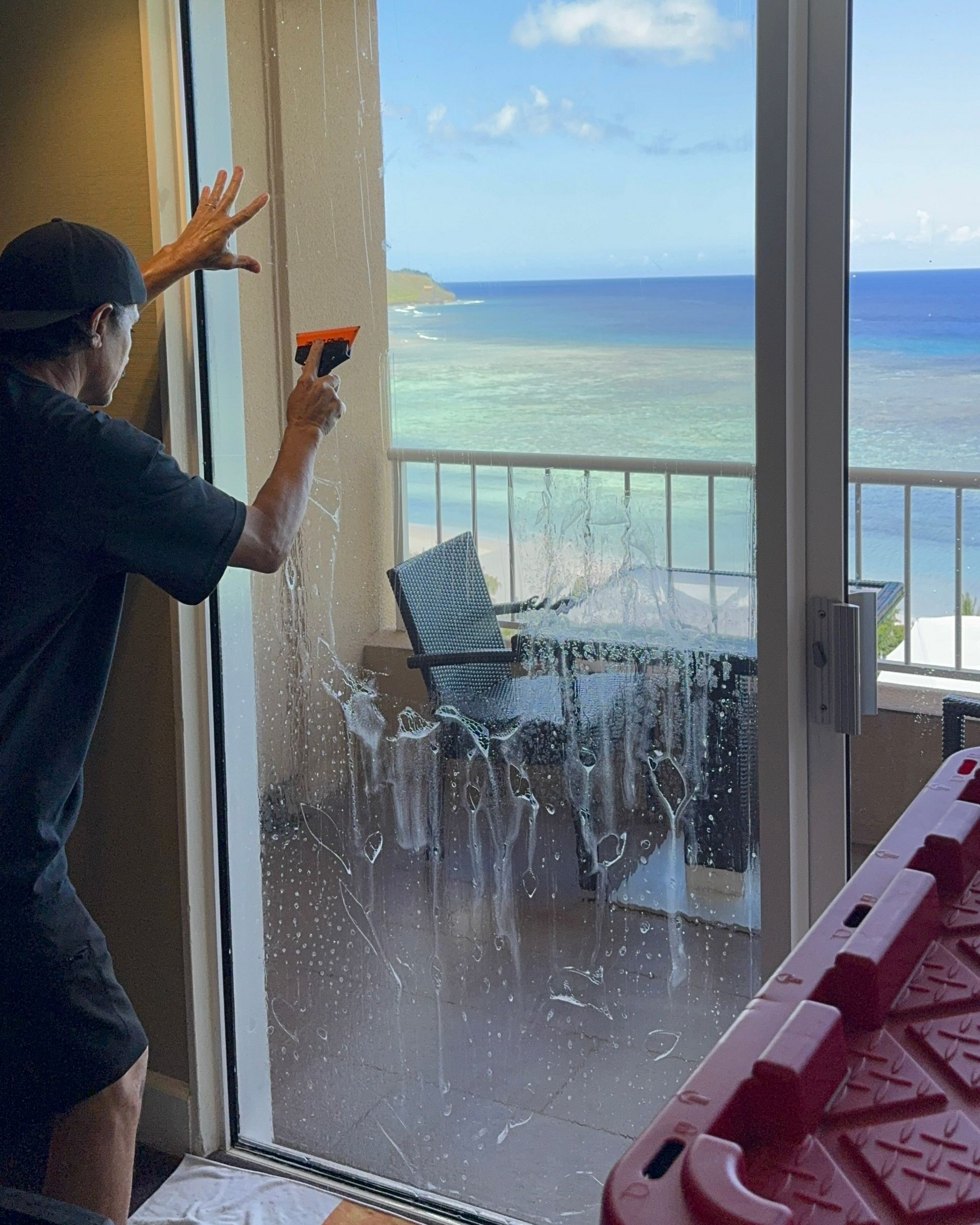 A person squeegees soapy water off a glass sliding door overlooking a scenic ocean balcony.