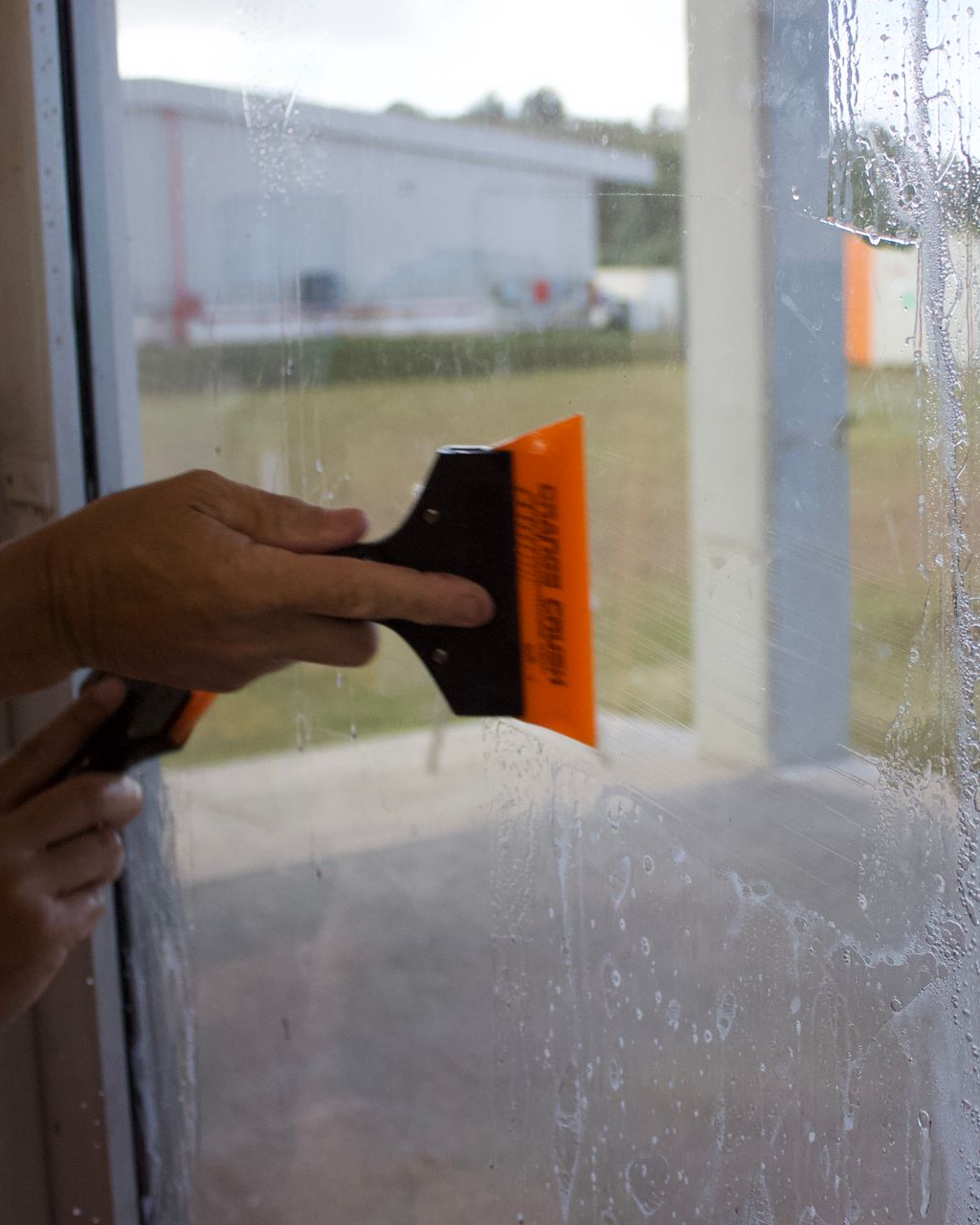 A person uses an orange-bladed squeegee to smooth a wet film onto a glass window, pushing out water and bubbles.