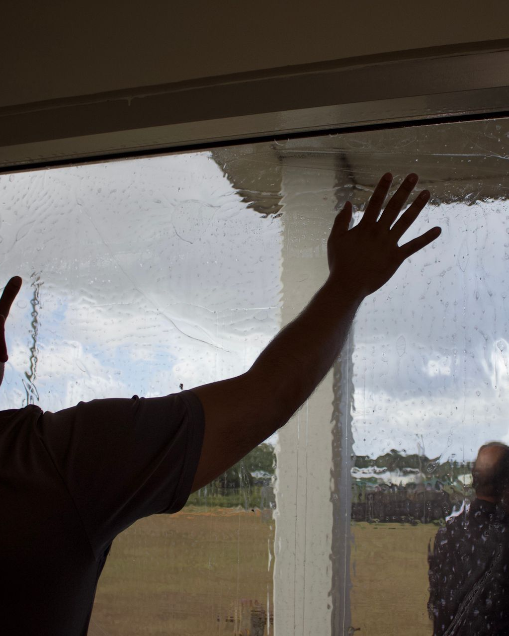 A person presses their hand against a rain-streaked window overlooking a field, with a glimpse of another person nearby.