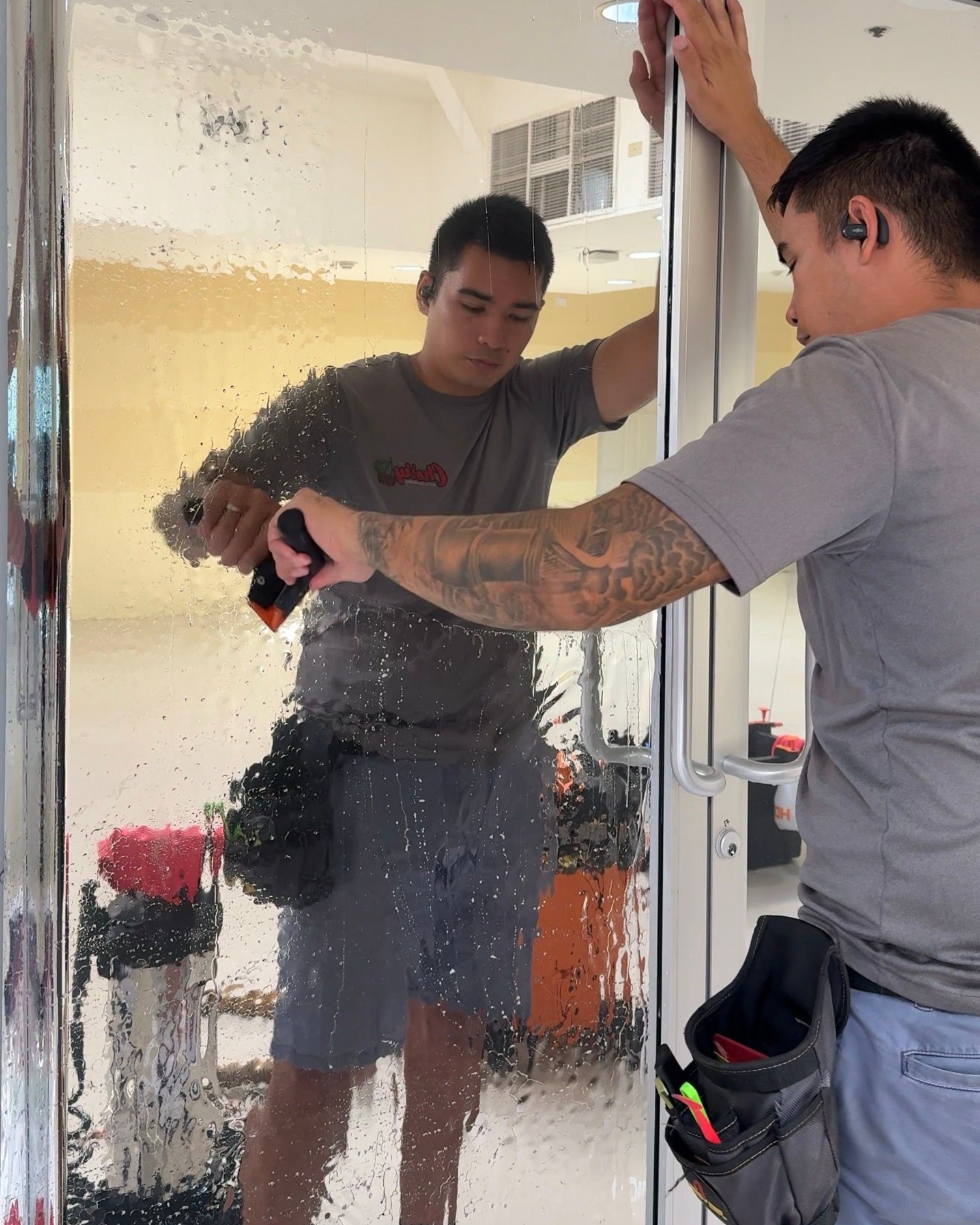 A person cleans a large glass pane with a squeegee, wearing a tool belt and a gray t-shirt.
