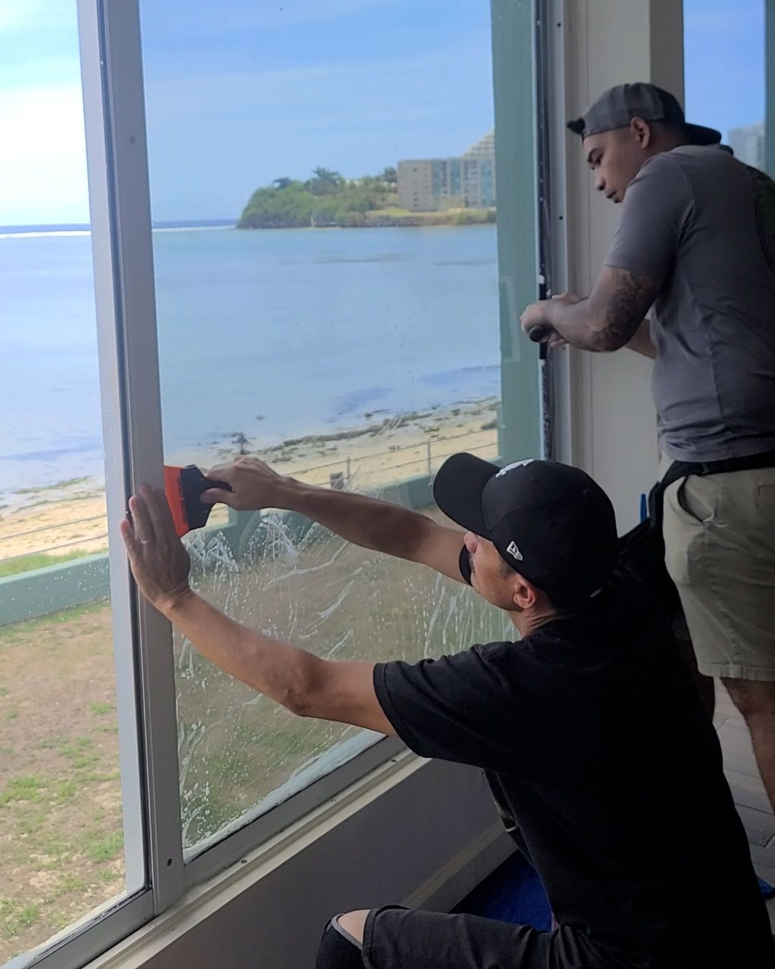 Two people work to install tint on a large window overlooking a scenic coastal view.