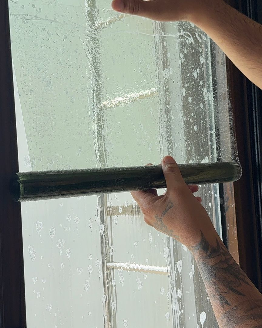 A person’s hands use a squeegee to smooth a wet layer of window tint onto a glass surface.