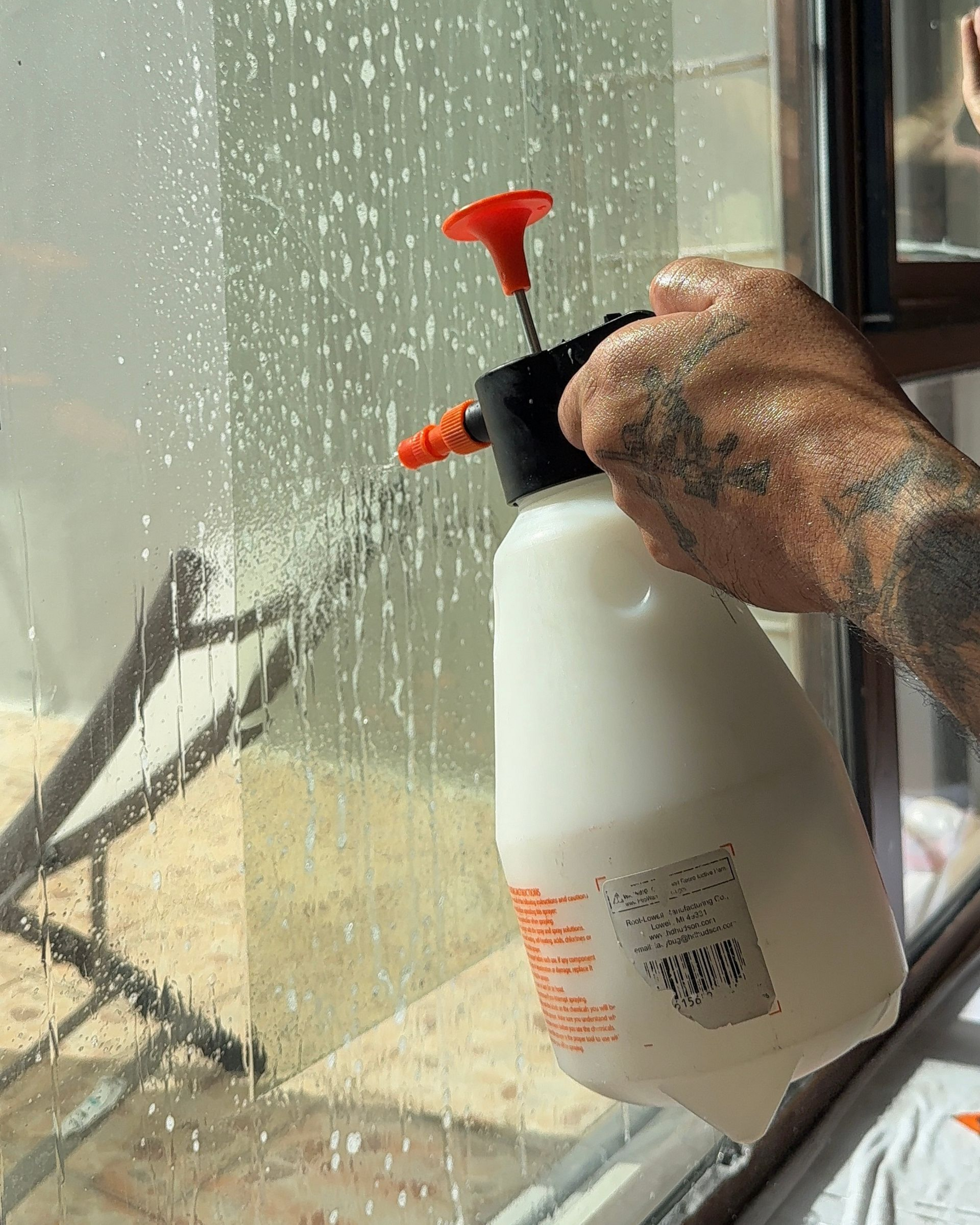 A tattooed hand holds a white pressurized spray bottle, misting a clear glass panel.