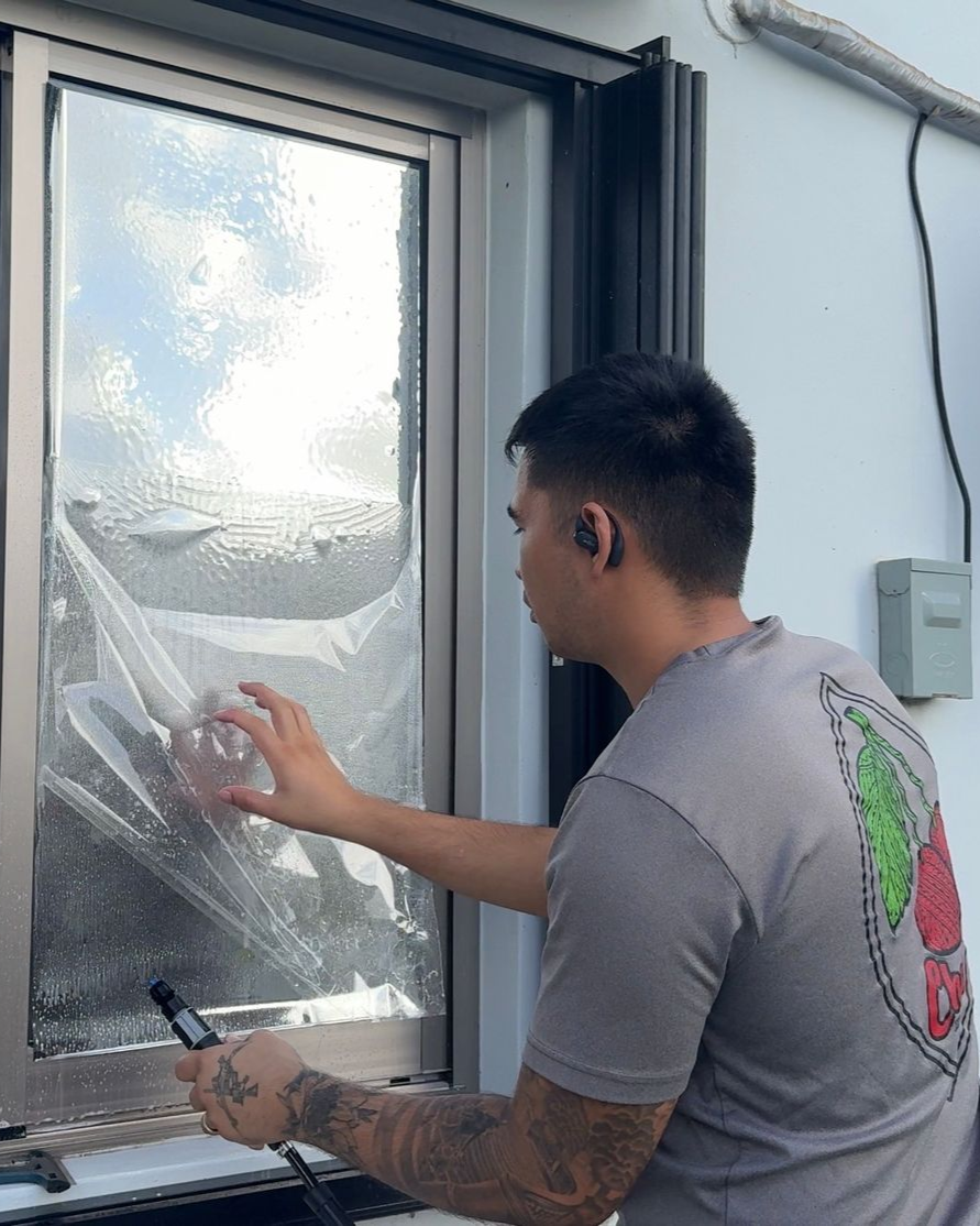 A person applies a reflective window film to a glass pane while holding a spray bottle.