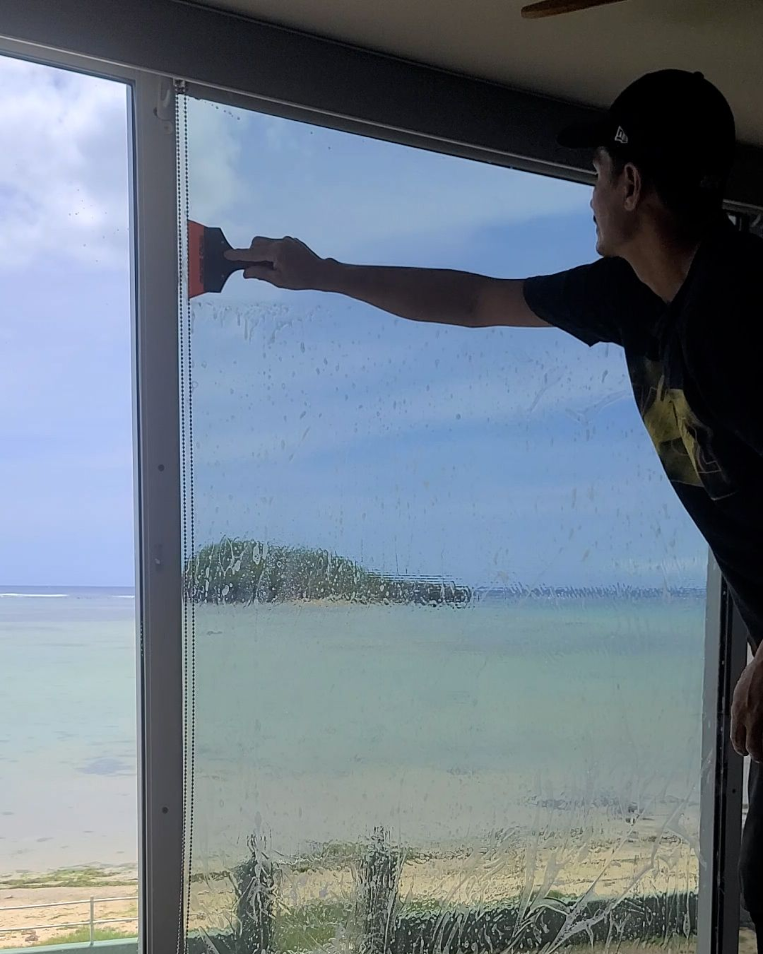 A person using a squeegee to apply window tint to a large glass pane overlooking a tropical beach and small island.
