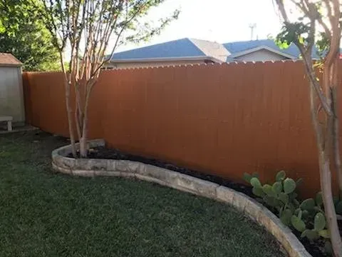 Brown privacy fence in a yard, bordered by a stone garden bed with grass, trees and cacti.