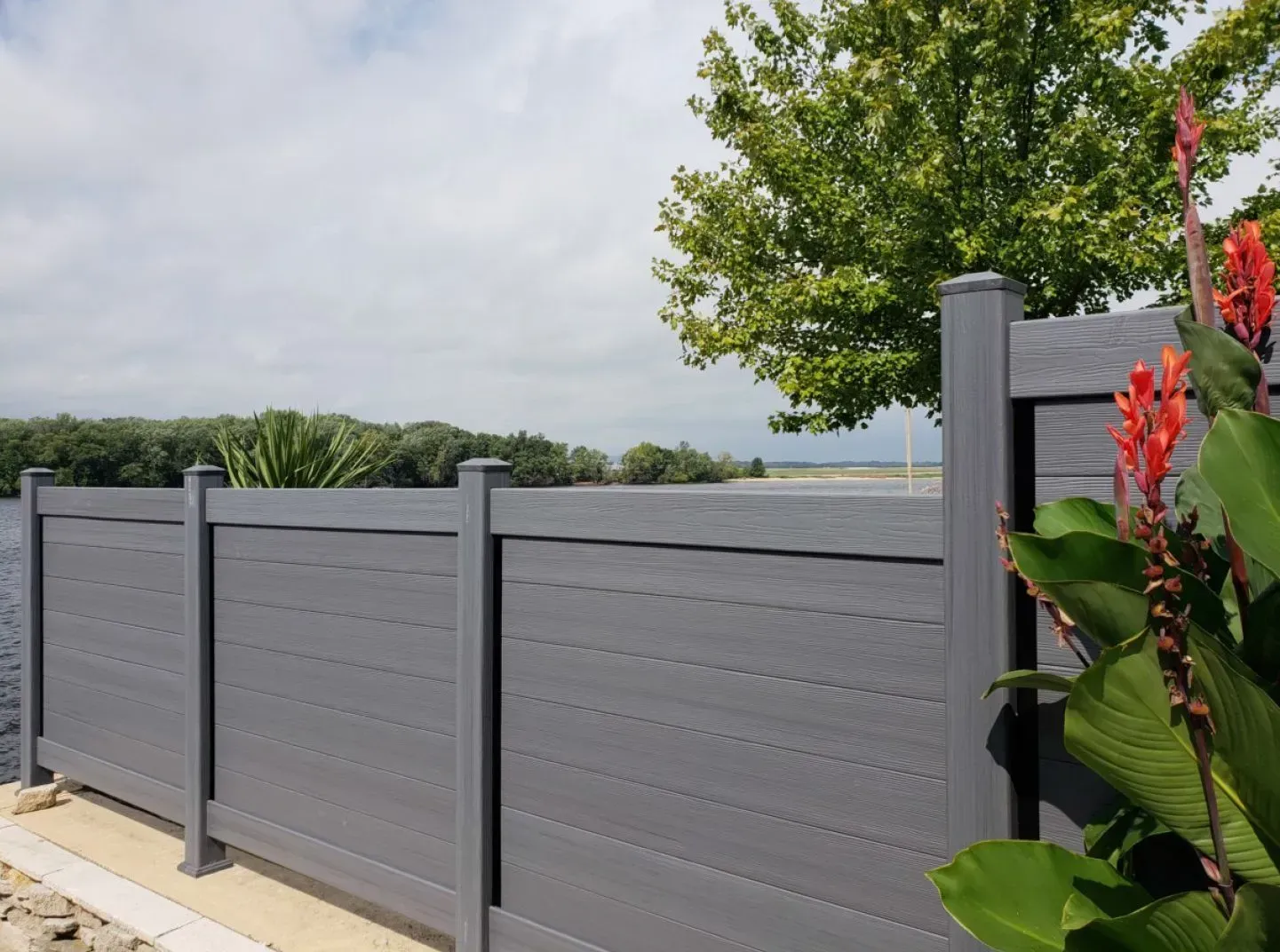 Gray composite fence by water, with red flowers in foreground and trees in the distance.