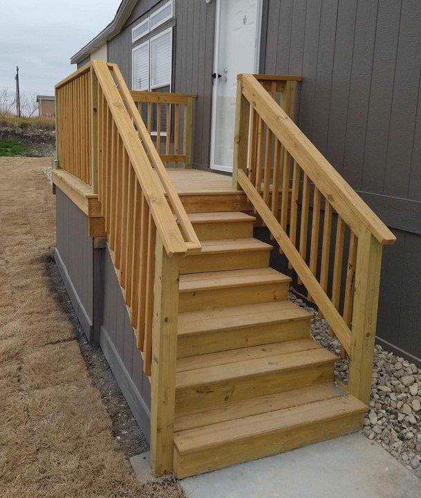 Wooden staircase leading to a gray house door with handrails, on a gravel and grass yard.