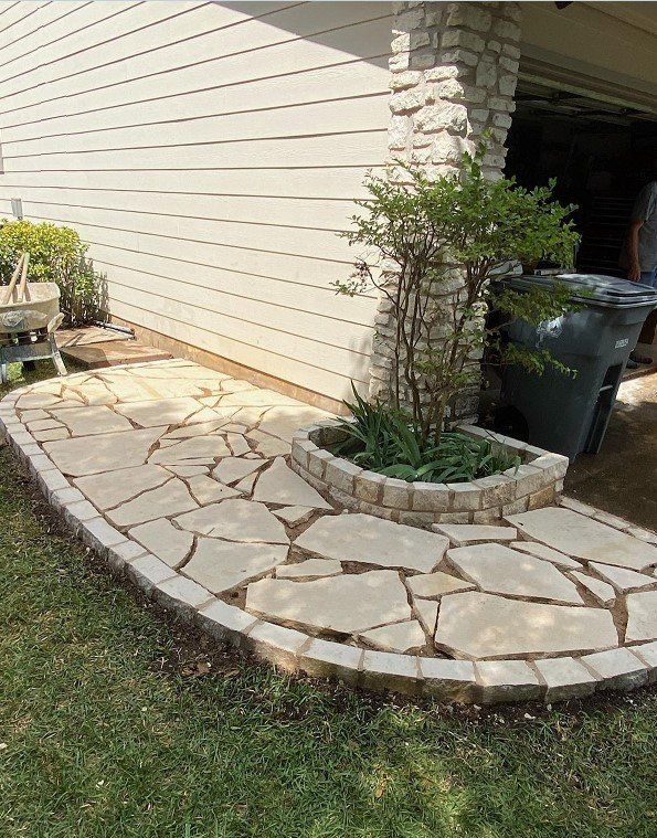 Stone path and flower bed next to a house. Green grass, beige stone, and a gray trash bin are visible.