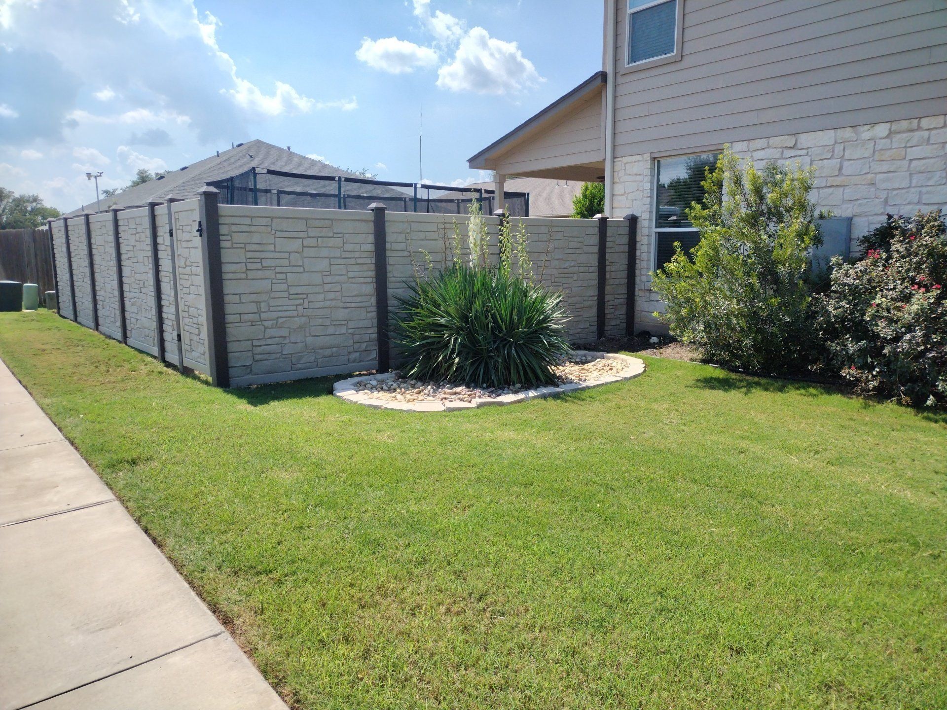 Gray and black patterned fence borders a green lawn and sidewalk next to a beige house.
