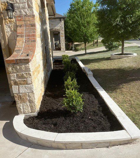 A stone planter with dark mulch and green shrubs borders a light stone wall, near a house.