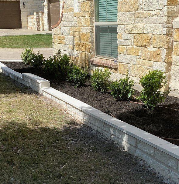 Stone-walled flower bed with mulch and green bushes in front of a house with stone siding.