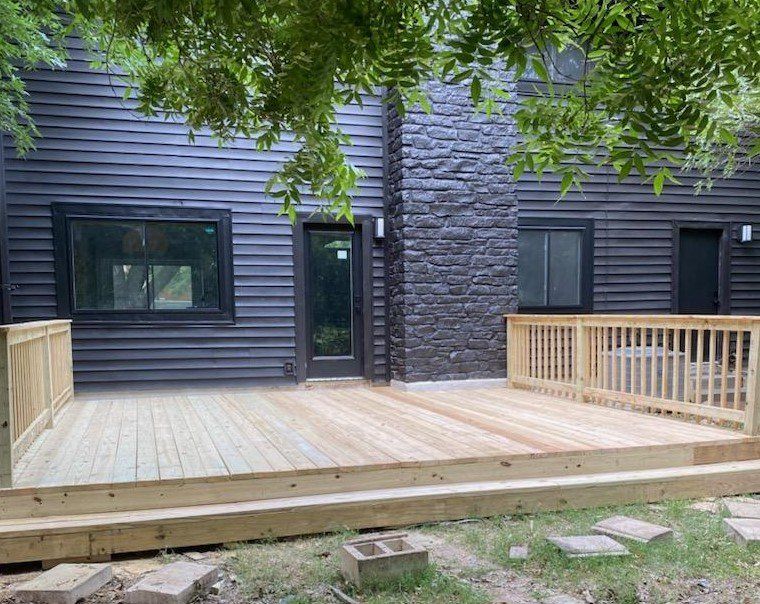 Newly built wooden deck attached to a dark gray house with black trimmed windows and a stone chimney.