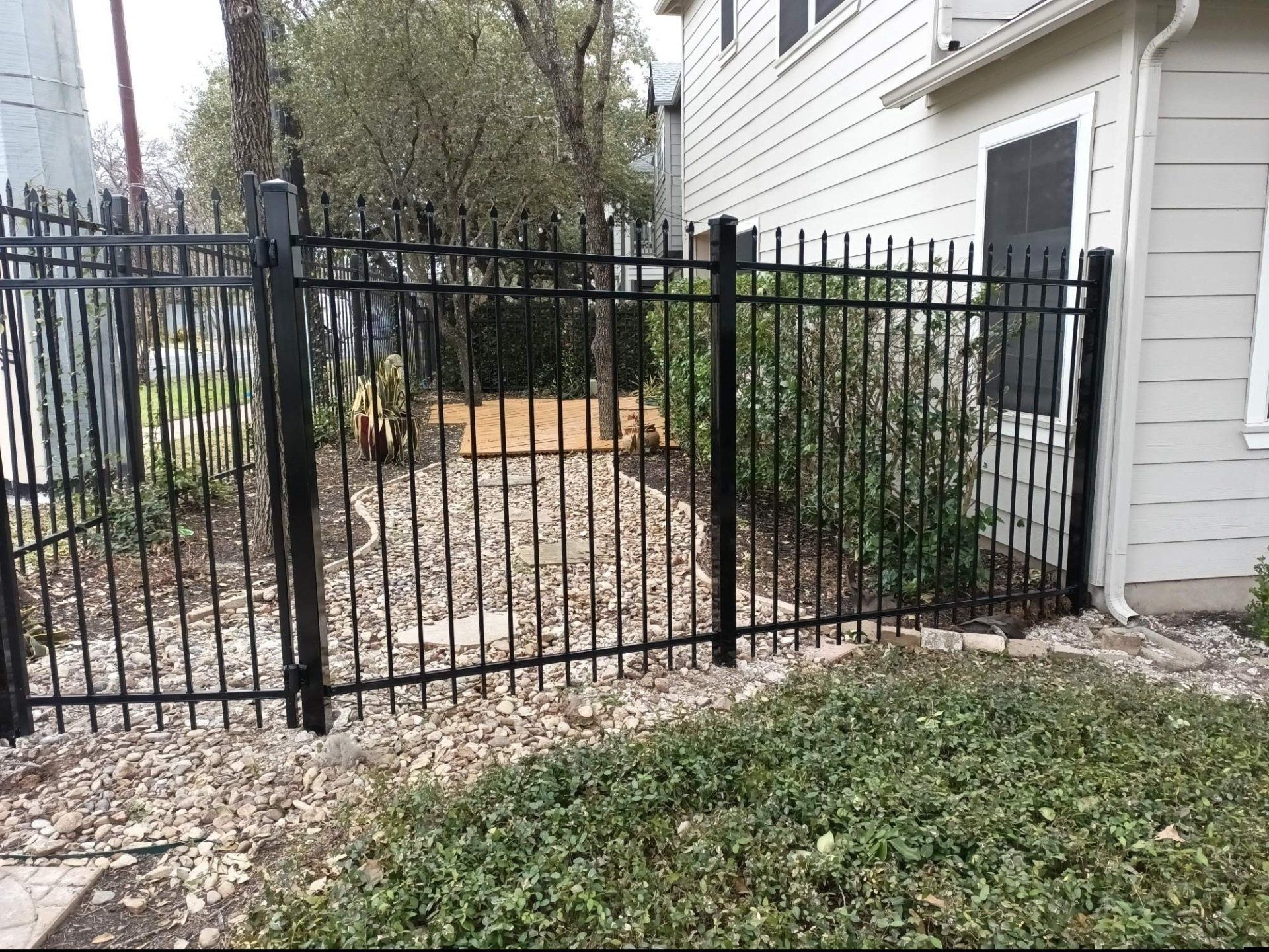 Black metal fence bordering a backyard with gravel and green bushes next to a light-colored house.