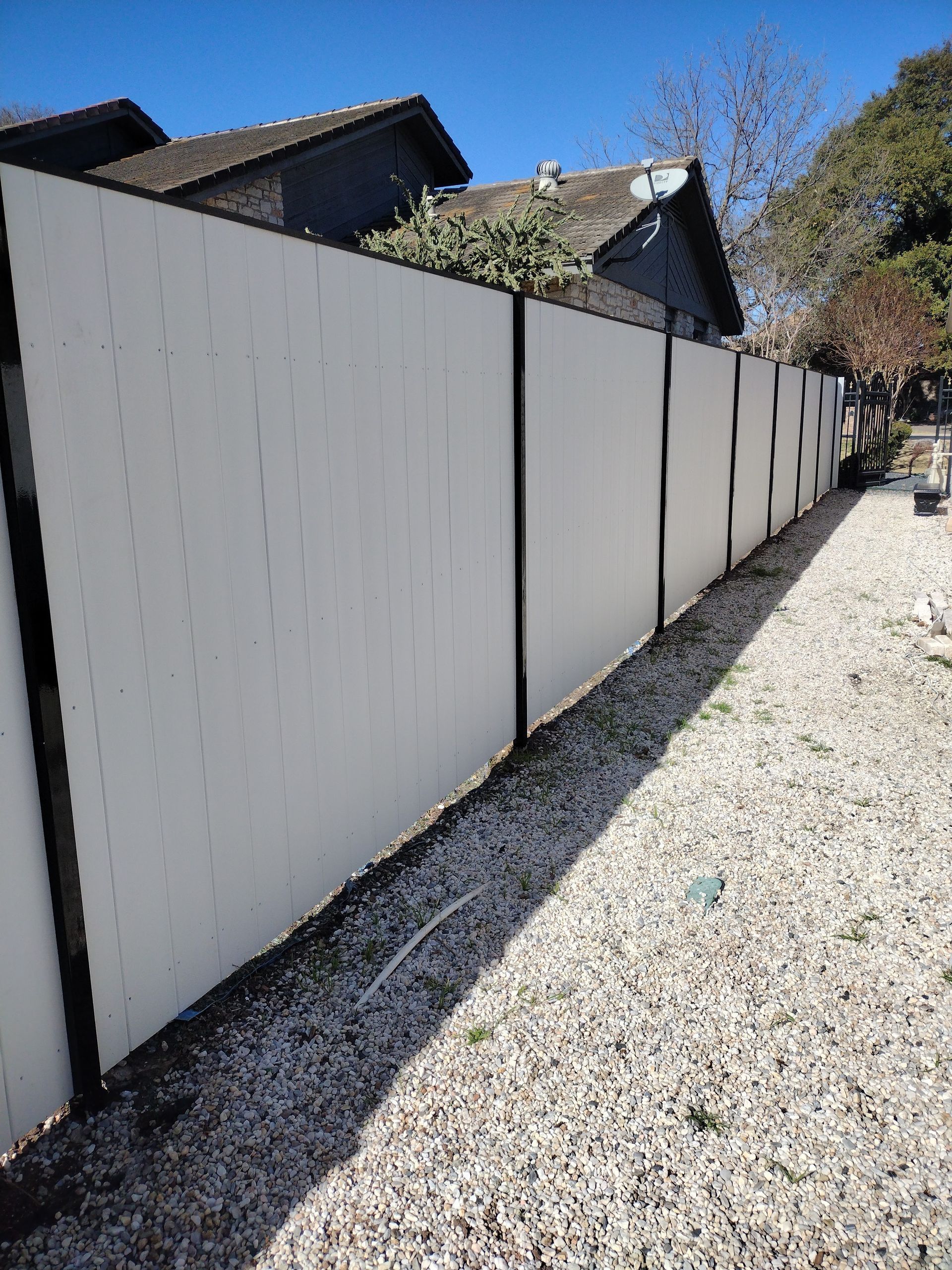 White privacy fence with black posts along a gravel path, trees and a house in the background.