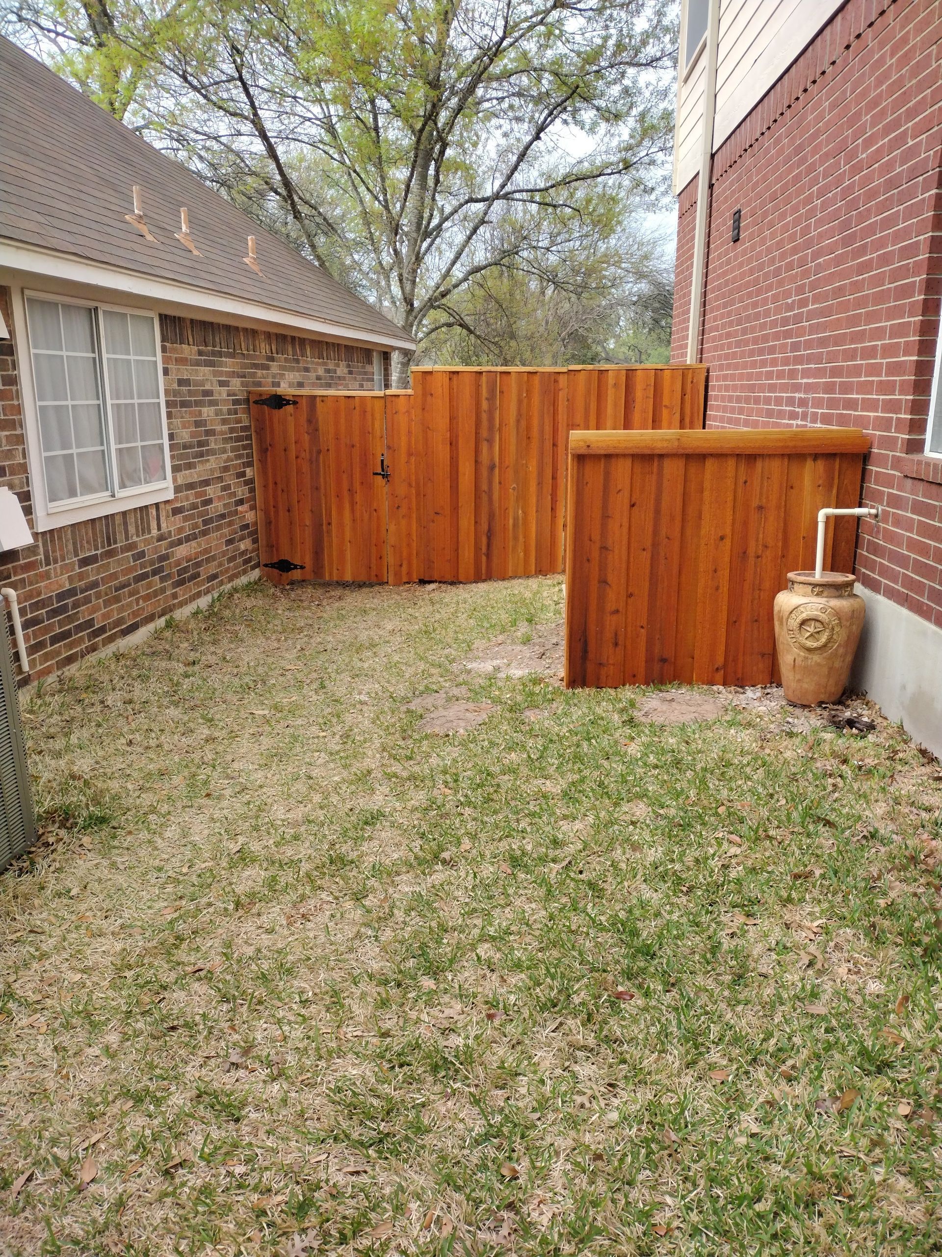 Wooden fence with brown stain surrounds a small backyard space, next to a brick house.