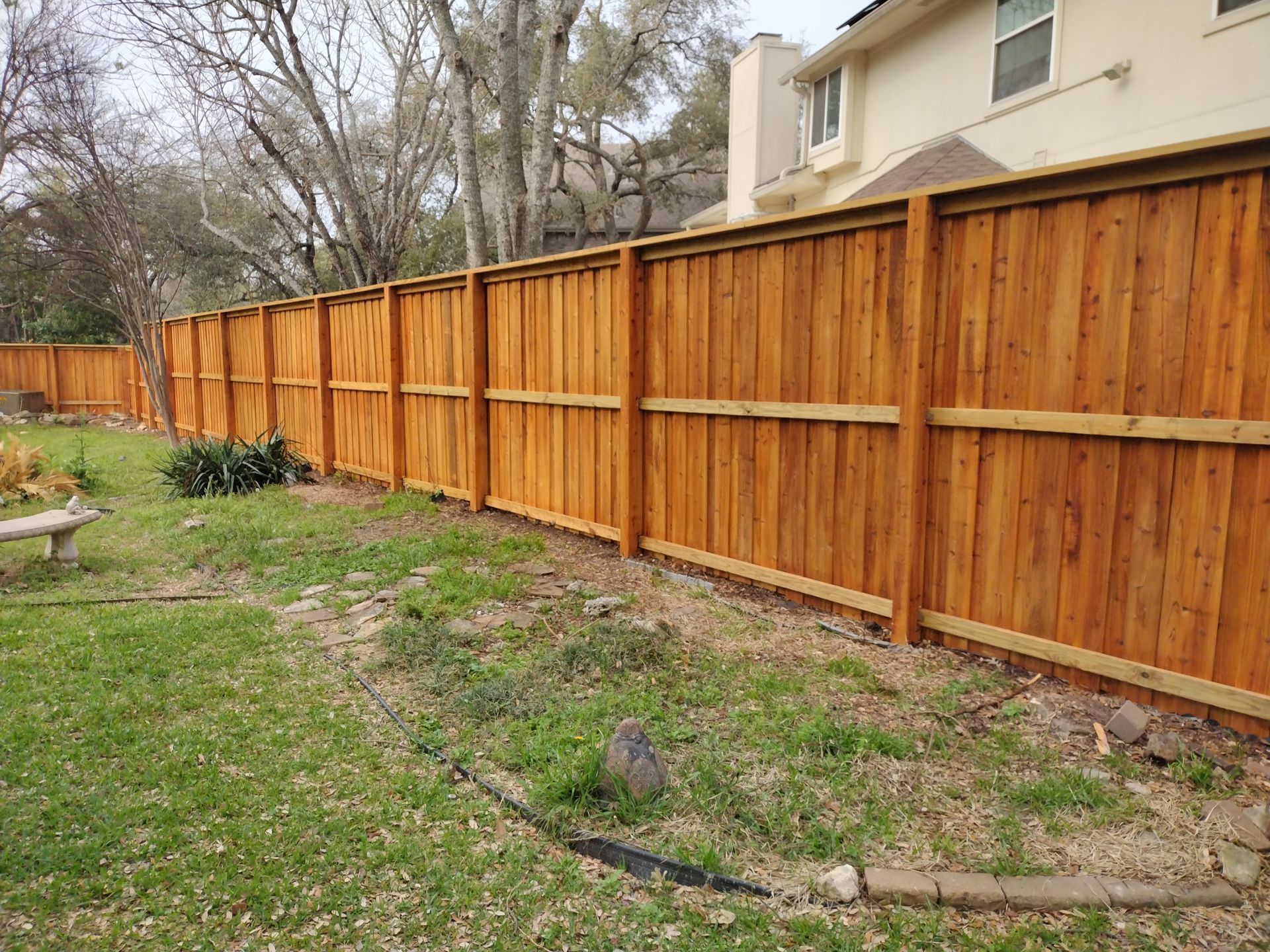 Wooden privacy fence in a backyard, stained brown, with green grass and trees in the background.