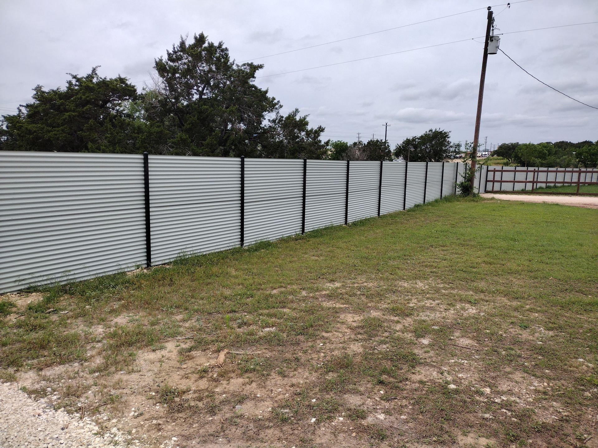 Corrugated metal fence with black posts on a grassy lot under a cloudy sky.