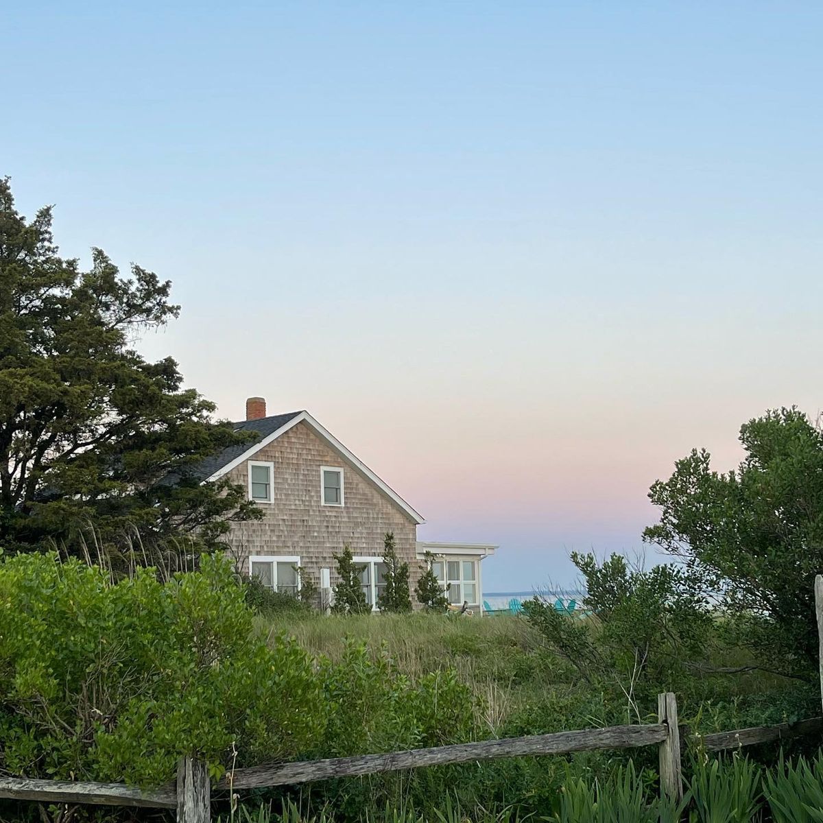Stone house overlooking a grassy dune at dusk, with trees and a pale pink sky over the sea