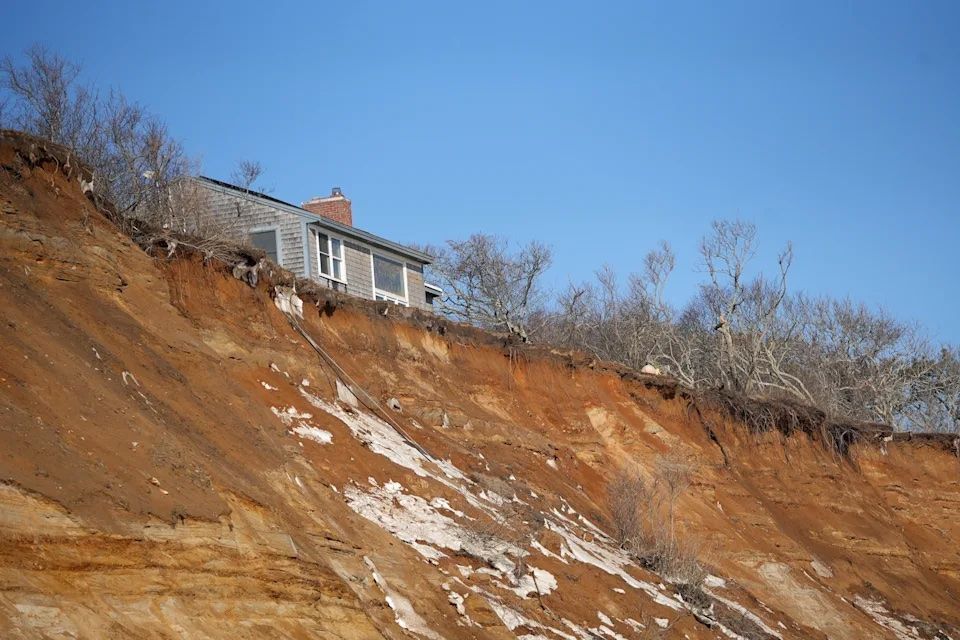 A house perched on the edge of a steep, eroding coastal cliff under a clear blue sky.