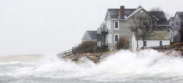 Large, churning ocean waves crash against the rocky shore near a seaside house during a storm.