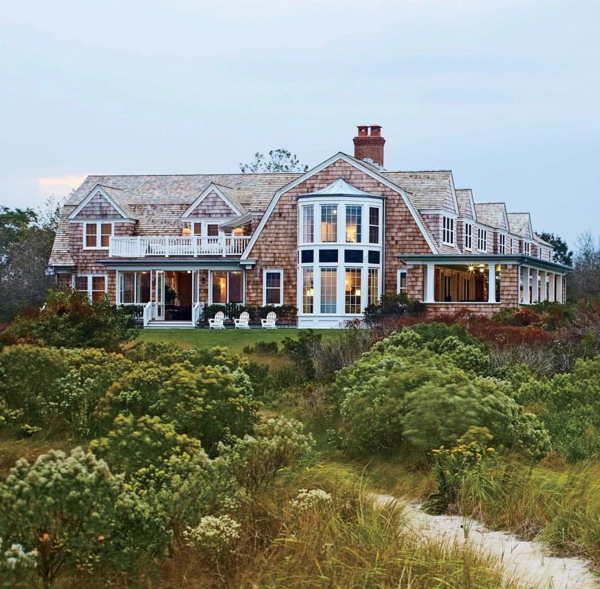 Large, weathered, multi-story shingle-clad house with multiple balconies and a covered porch in a grassy dune setting.