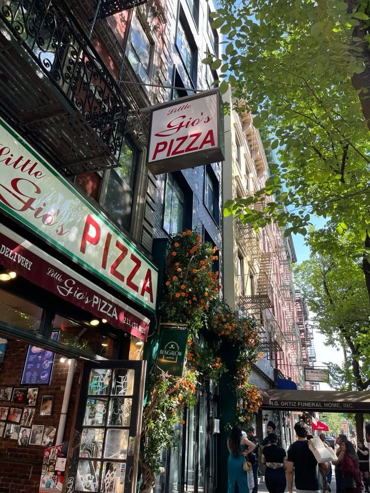 Street view of Little Gio’s Pizza storefront with bright red signs, flowers, and people walking by.