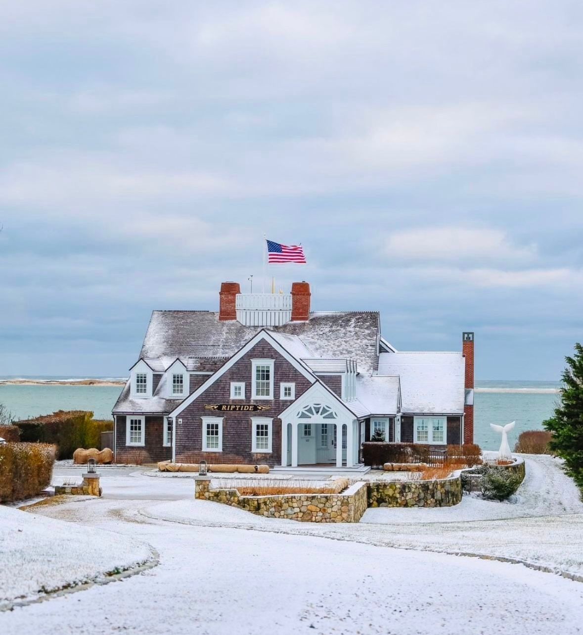 Snow-covered house with American flag on rooftop. Ocean in the background. Cloudy sky.
