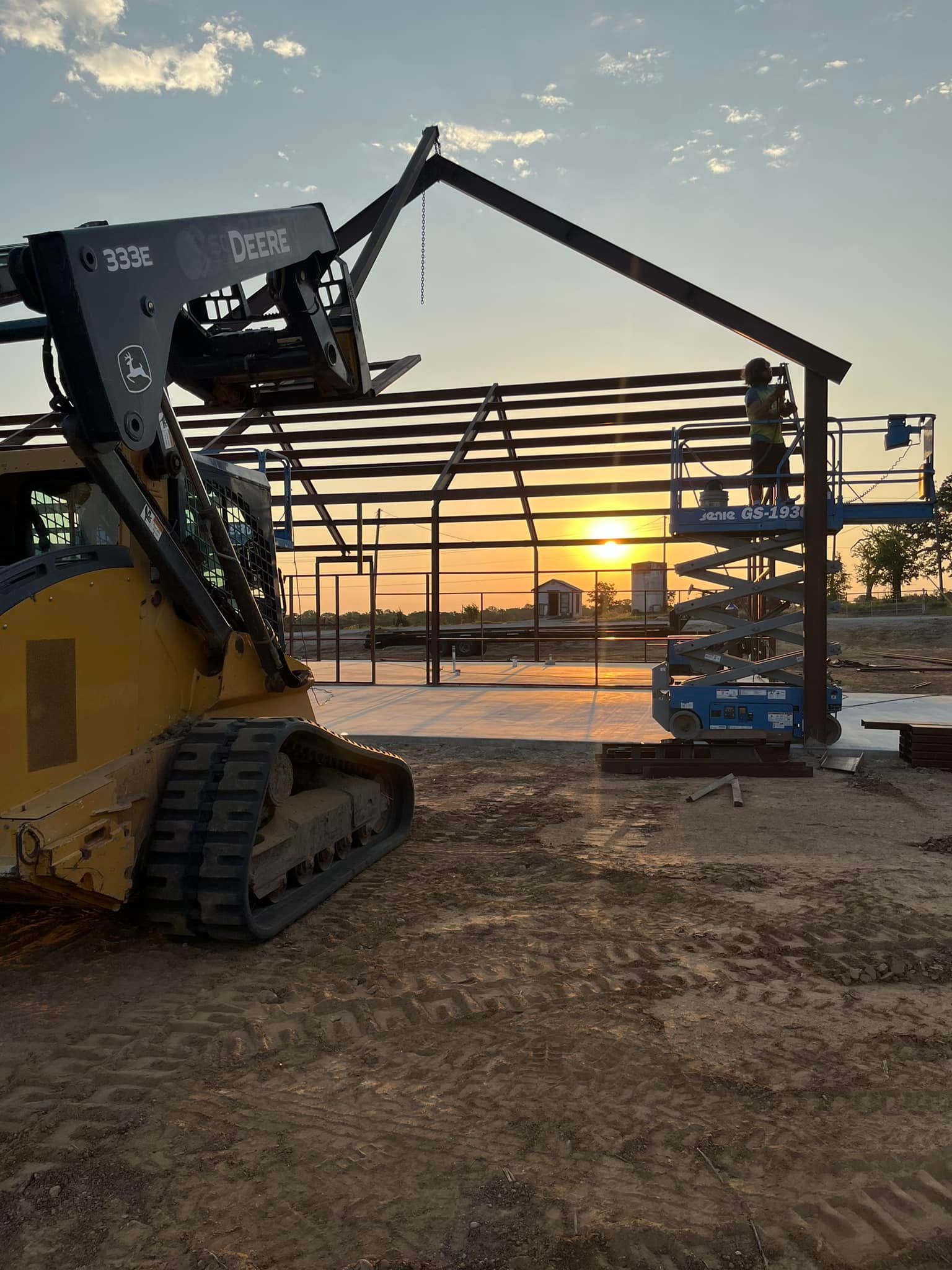 A bulldozer is lifting a metal structure on a construction site at sunset.