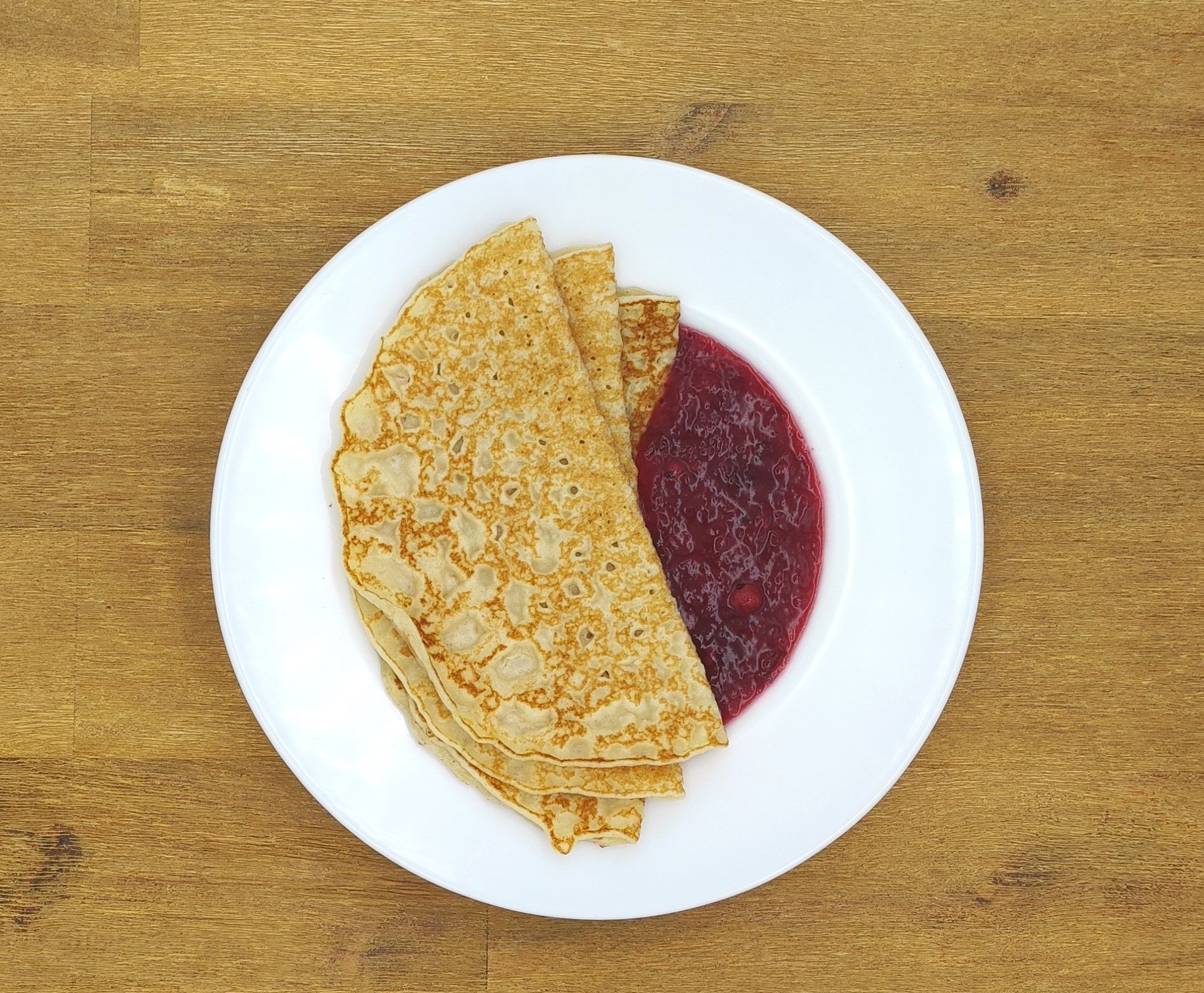 Ein Stapel Crêpes mit roter Marmelade auf einem weißen Teller, Holztisch.