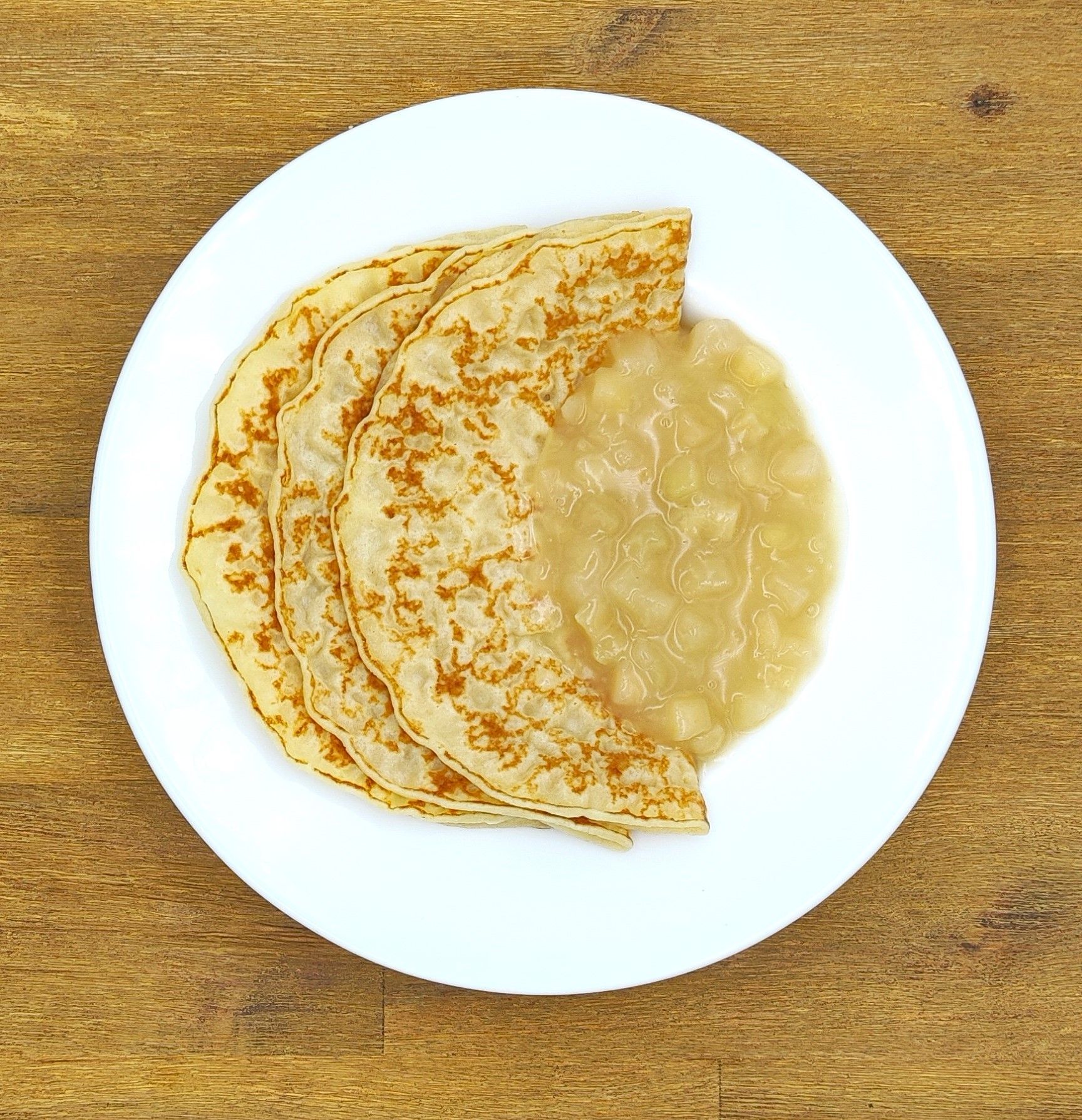 Drei goldgelbe Pfannkuchen auf einem weißen Teller mit Apfelmus, angerichtet auf einer Holzplatte.