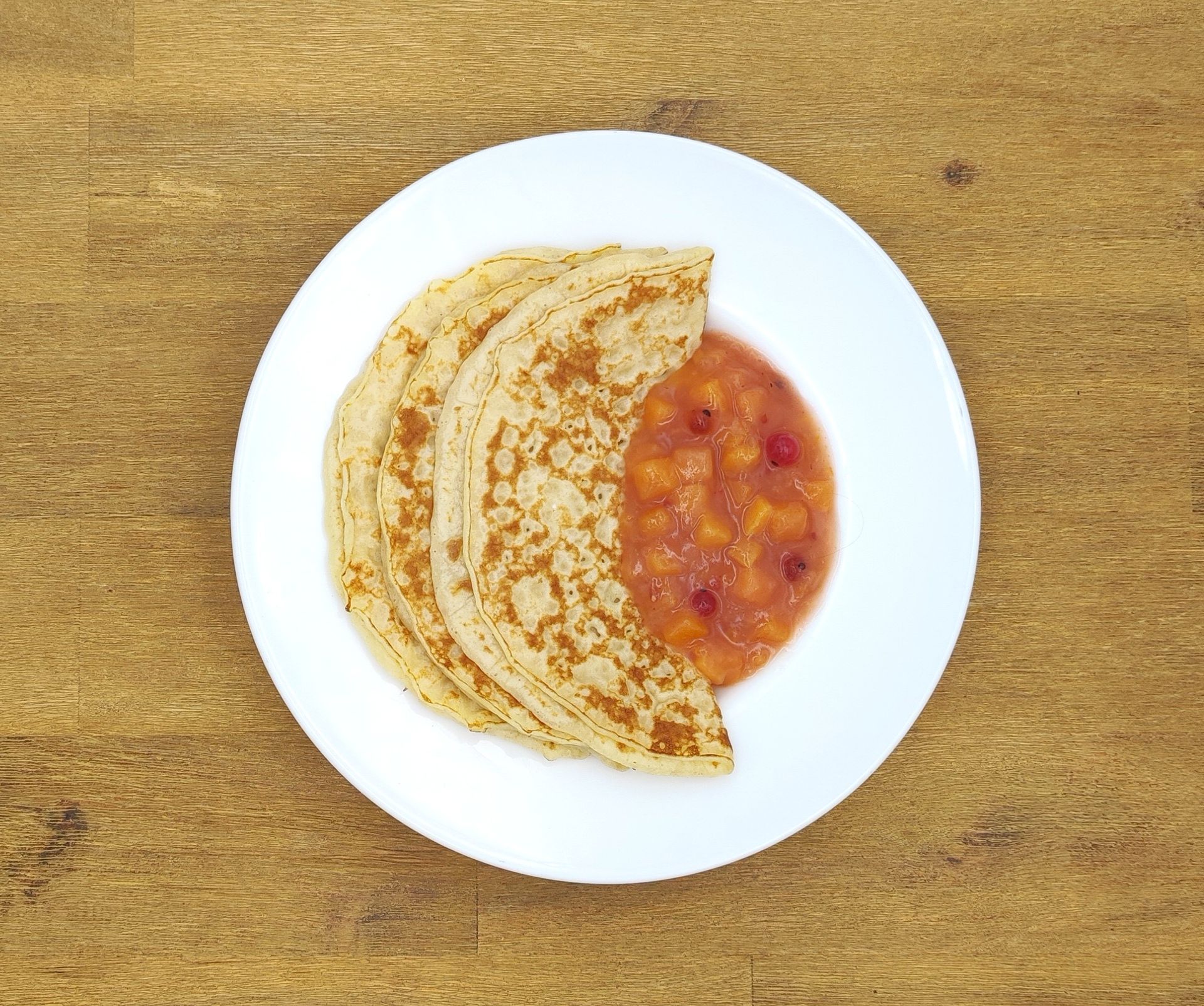 Drei Pfannkuchen mit Pfirsichkompott auf einem weißen Teller, angerichtet auf einem Holztisch.