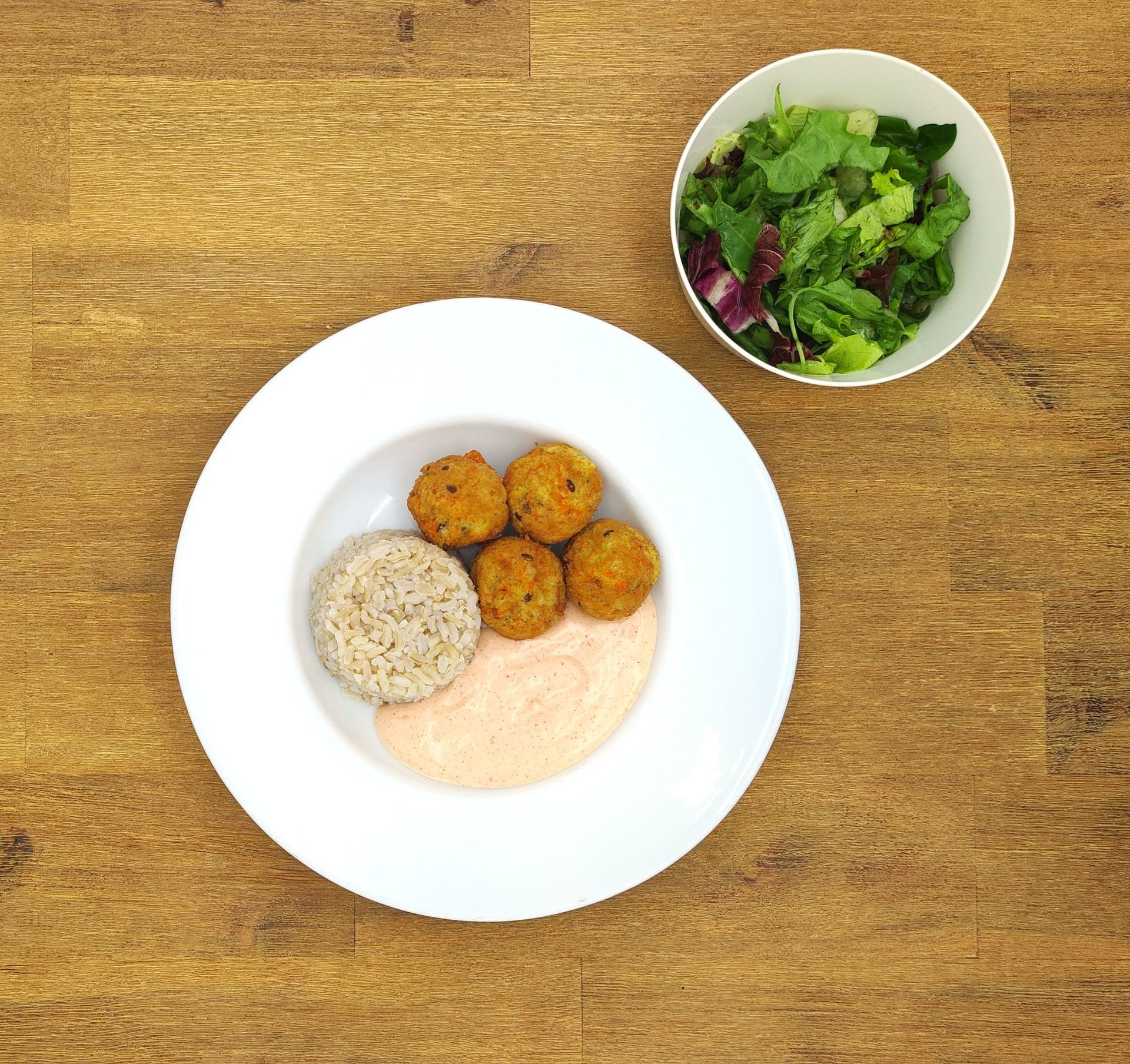 Ein Teller mit Falafel, Reis und Soße sowie eine Schüssel Salat auf einem Holztisch.