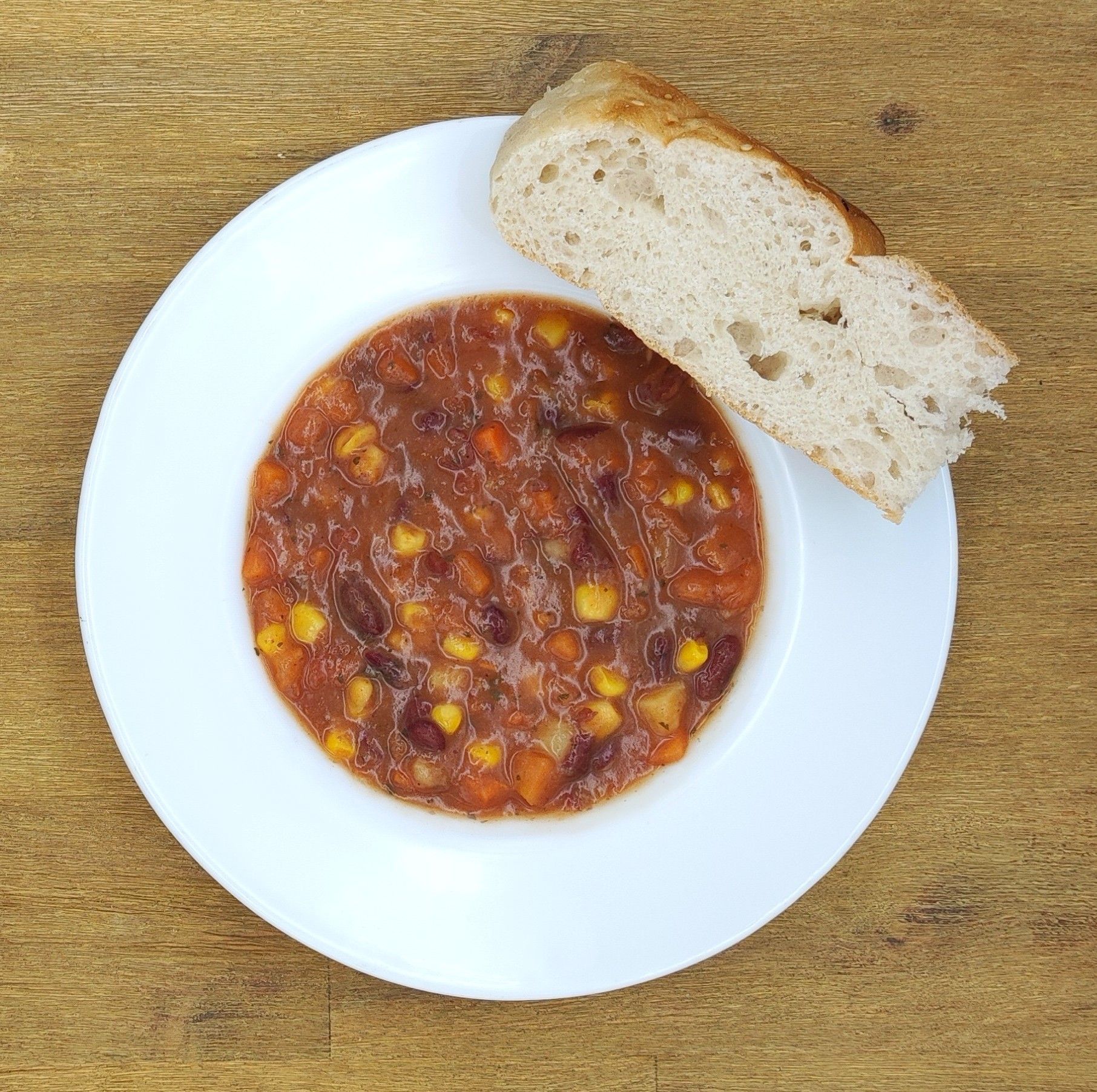Eine Schüssel Chili mit Mais, Bohnen und Karotten, serviert mit einer Scheibe Brot auf einem weißen Teller.