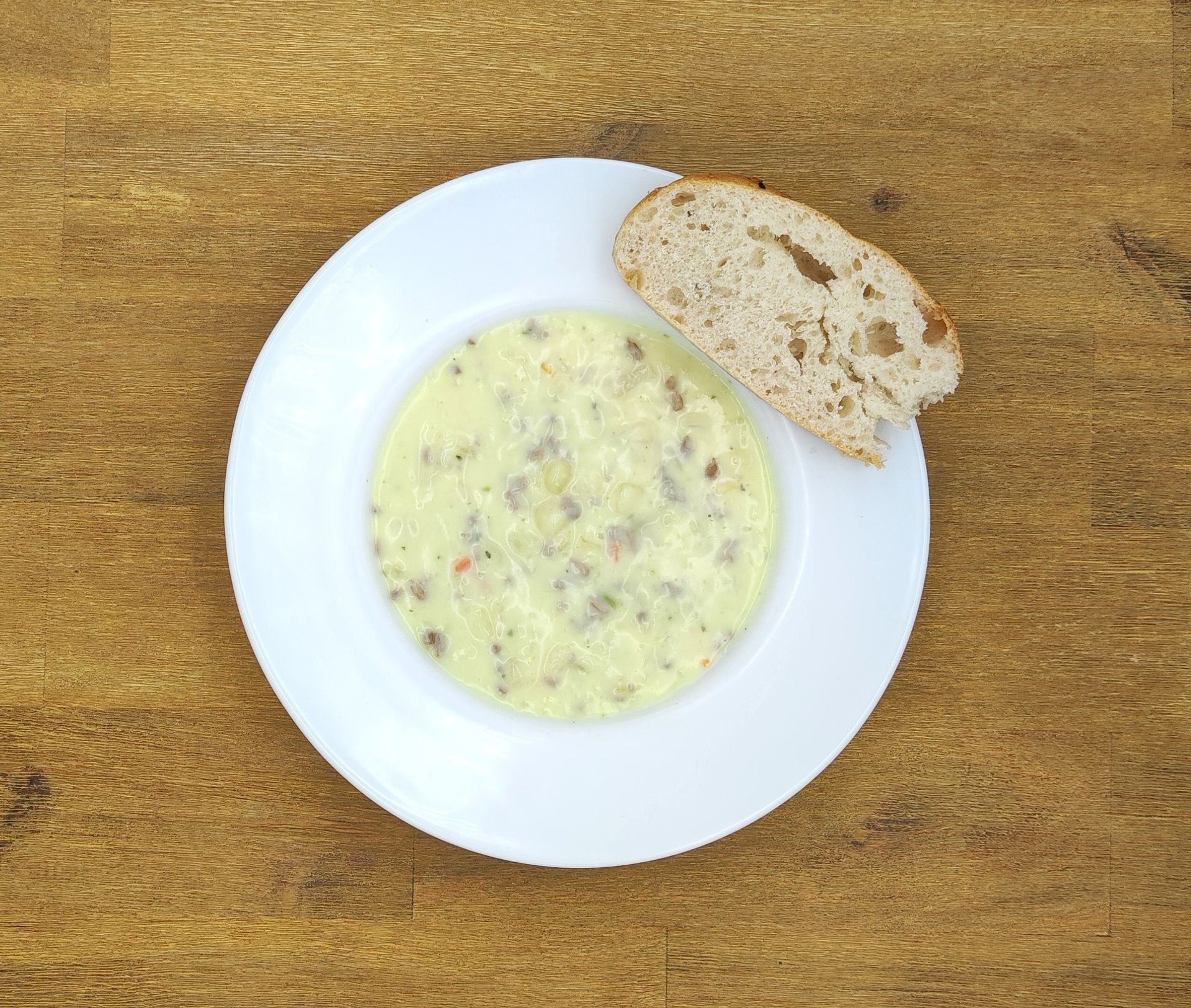Eine Schüssel cremige Suppe mit Fleischstücken, serviert mit einer Scheibe Brot auf einem weißen Teller auf einer Holzplatte.