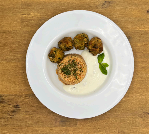 Ein weißer Teller mit frittierten Gemüsebällchen, Couscous, weißer Soße und einem Minzzweig auf einem Holztisch.