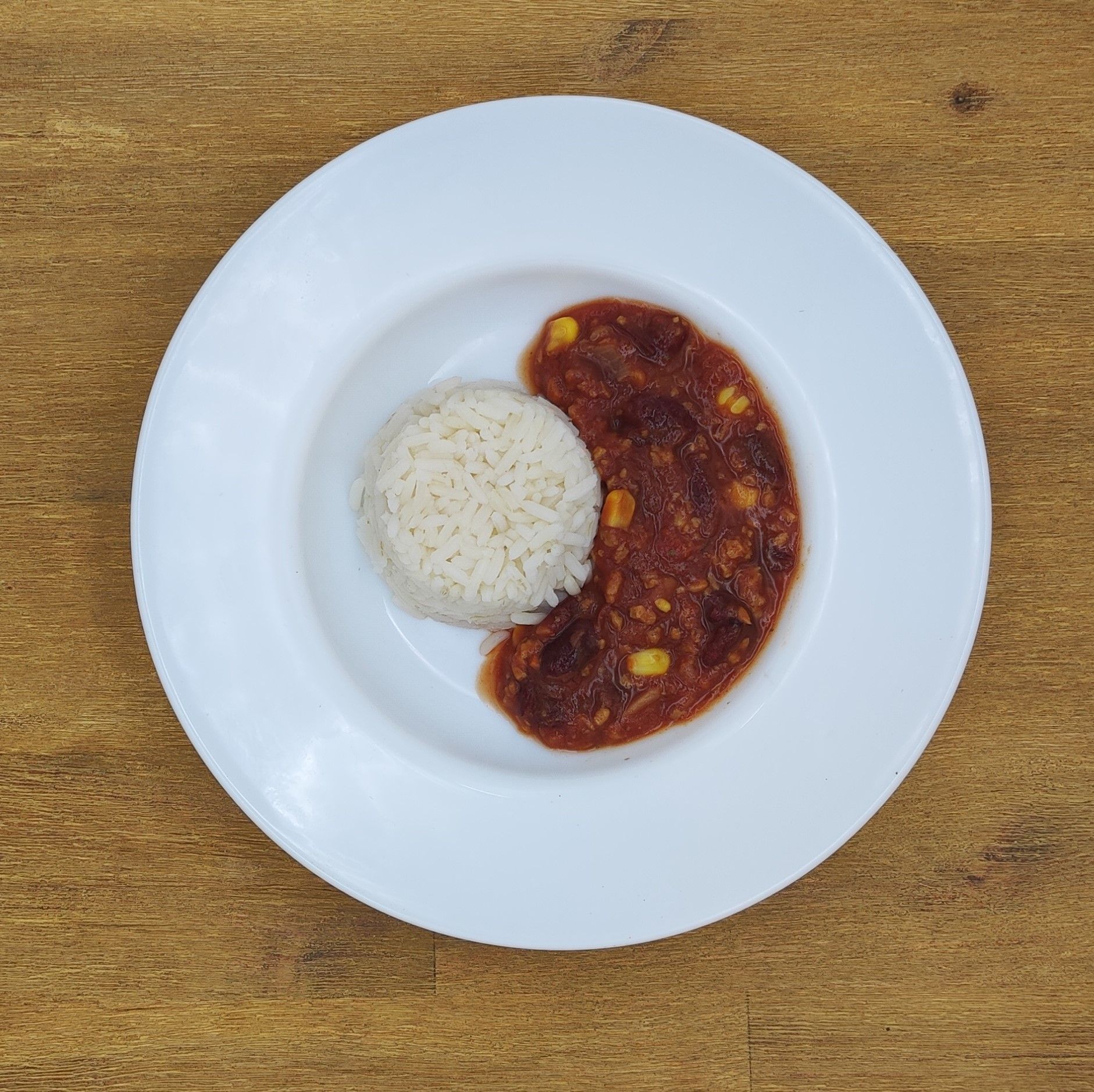 Weißer Teller mit Reis und rötlich-braunem Eintopf auf einem Holztisch.