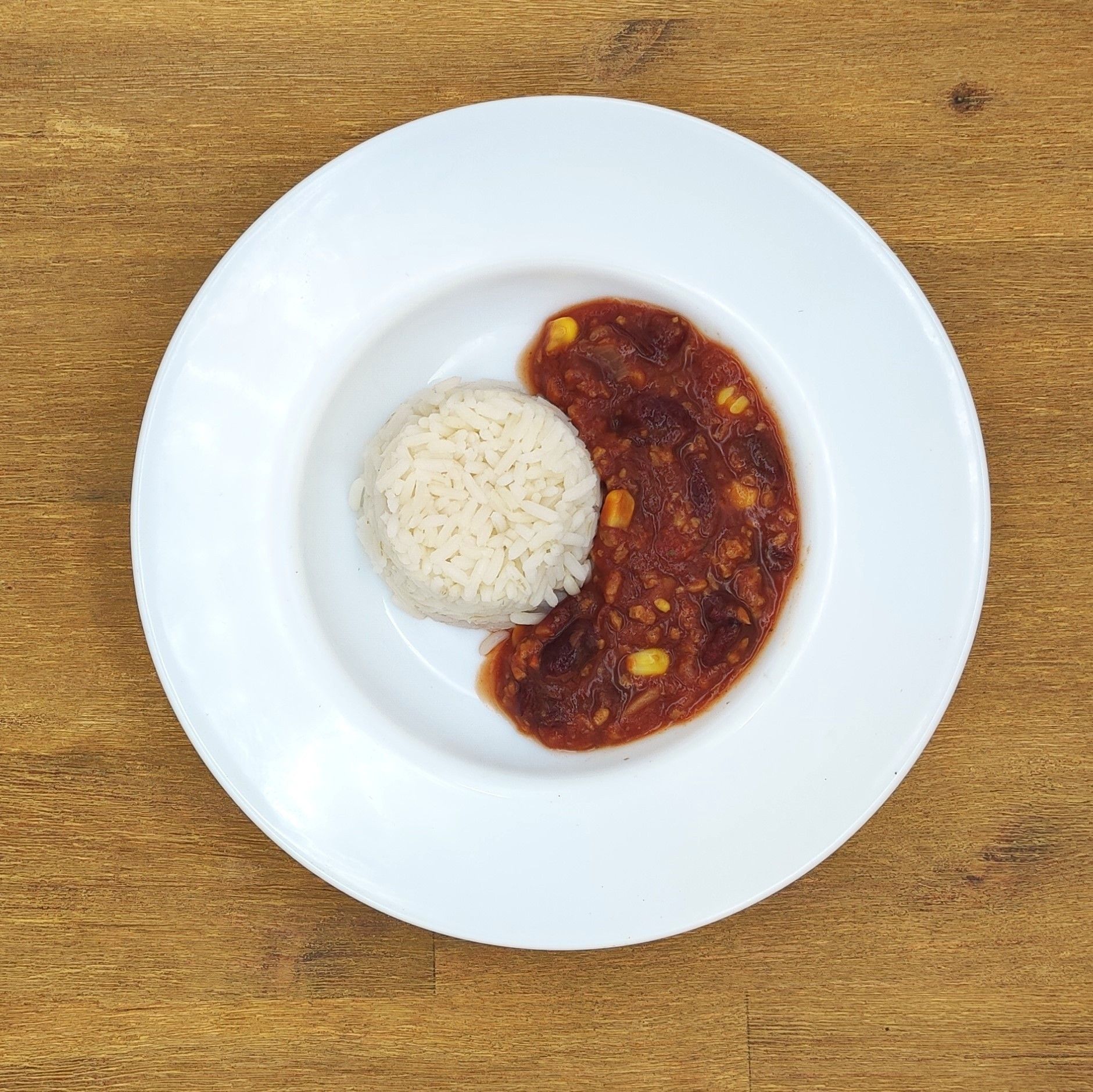 Weißer Teller mit Reis und Chili auf einem Holztisch.