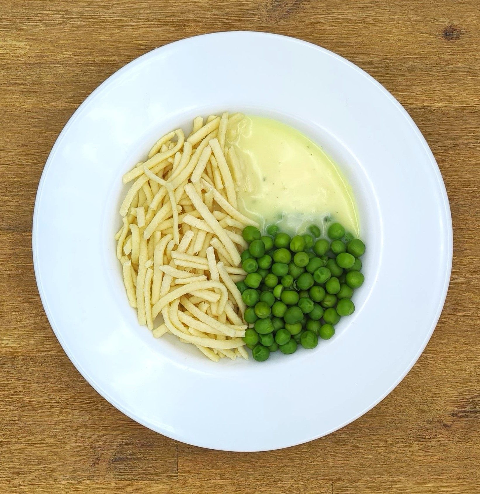 Weißer Teller mit Nudeln, grünen Erbsen und cremiger Soße auf einer Holzoberfläche.