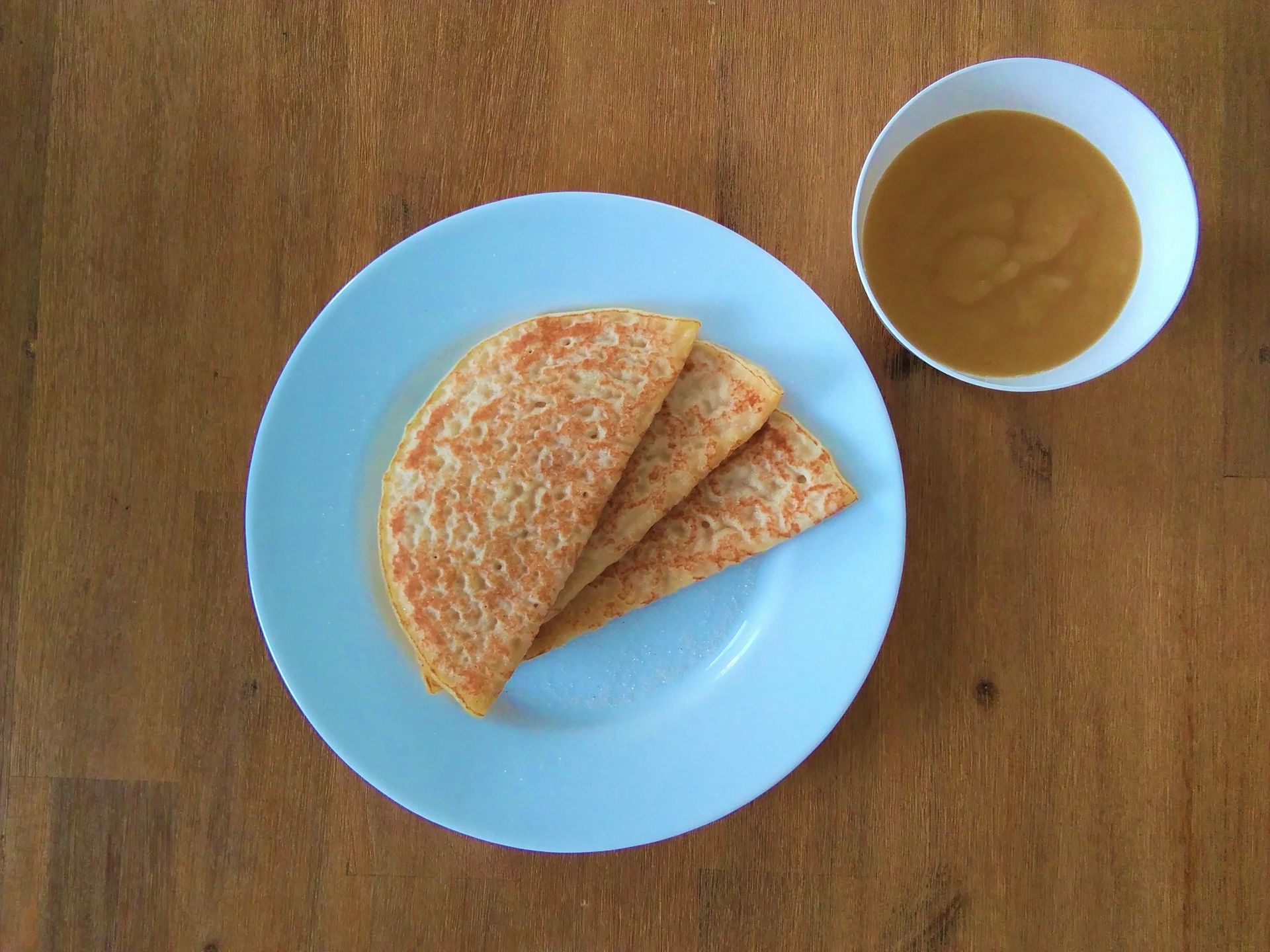 Drei gefaltete Crêpes auf einem blauen Teller, daneben eine Schüssel mit brauner Soße, auf einer Holzoberfläche.