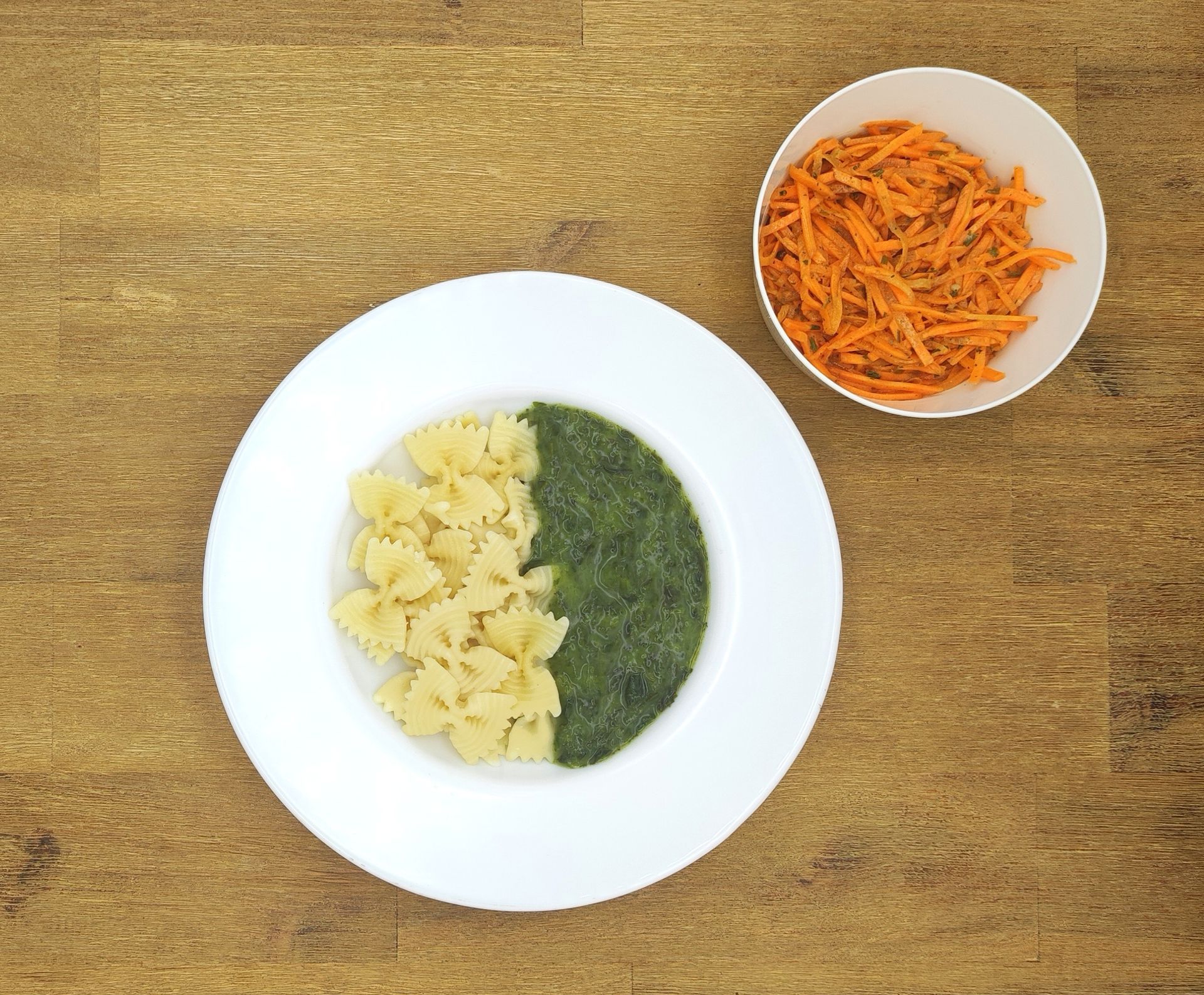 Pasta mit grüner Soße und geriebenen Karotten in weißen Schüsseln auf Holz.