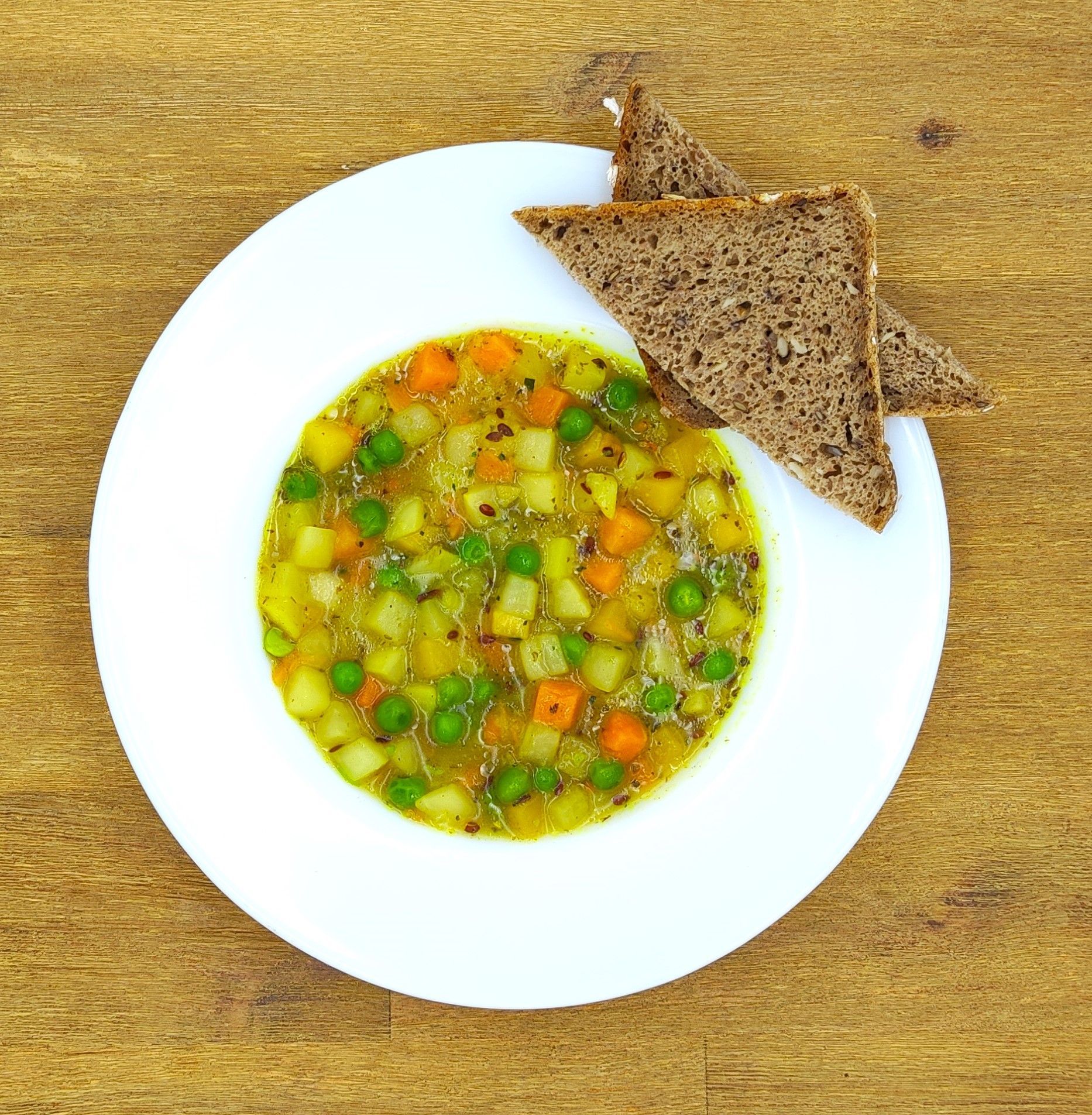 Gemüsesuppe mit Karotten, Erbsen und Kartoffeln auf einem weißen Teller mit dunklem Brot auf einem Holztisch.
