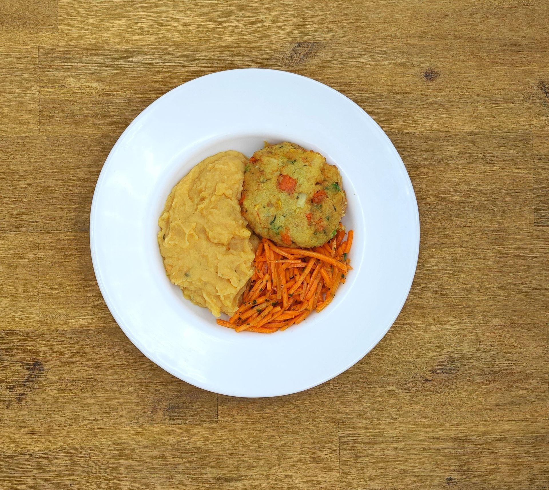 Teller mit einer gelben Schüssel, einem Gemüsebratling und geraspelten Karotten auf einem Holztisch.