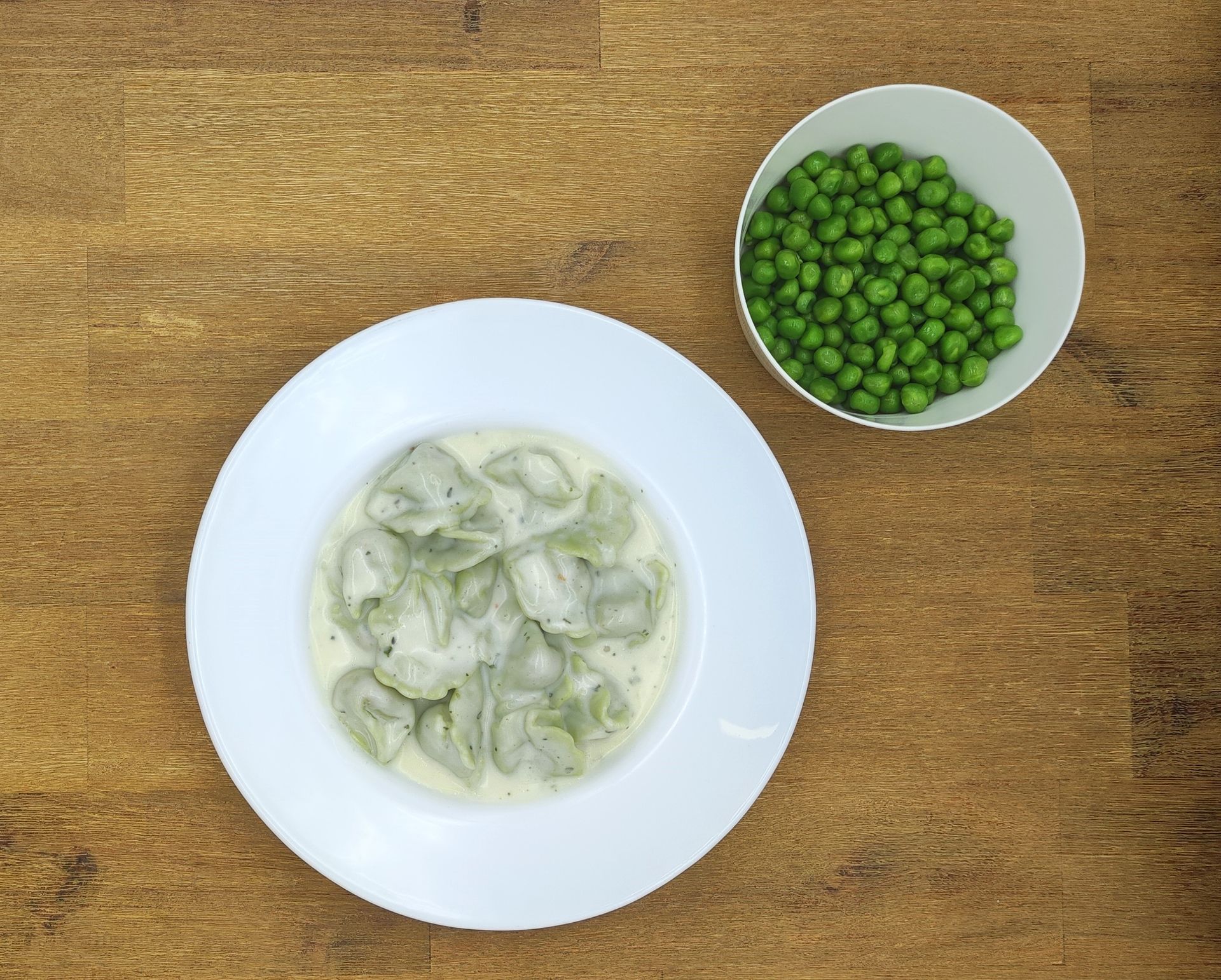 Ein weißer Teller mit grünen Tortellini in Sahnesauce, eine Schüssel mit Erbsen auf einem Holztisch.