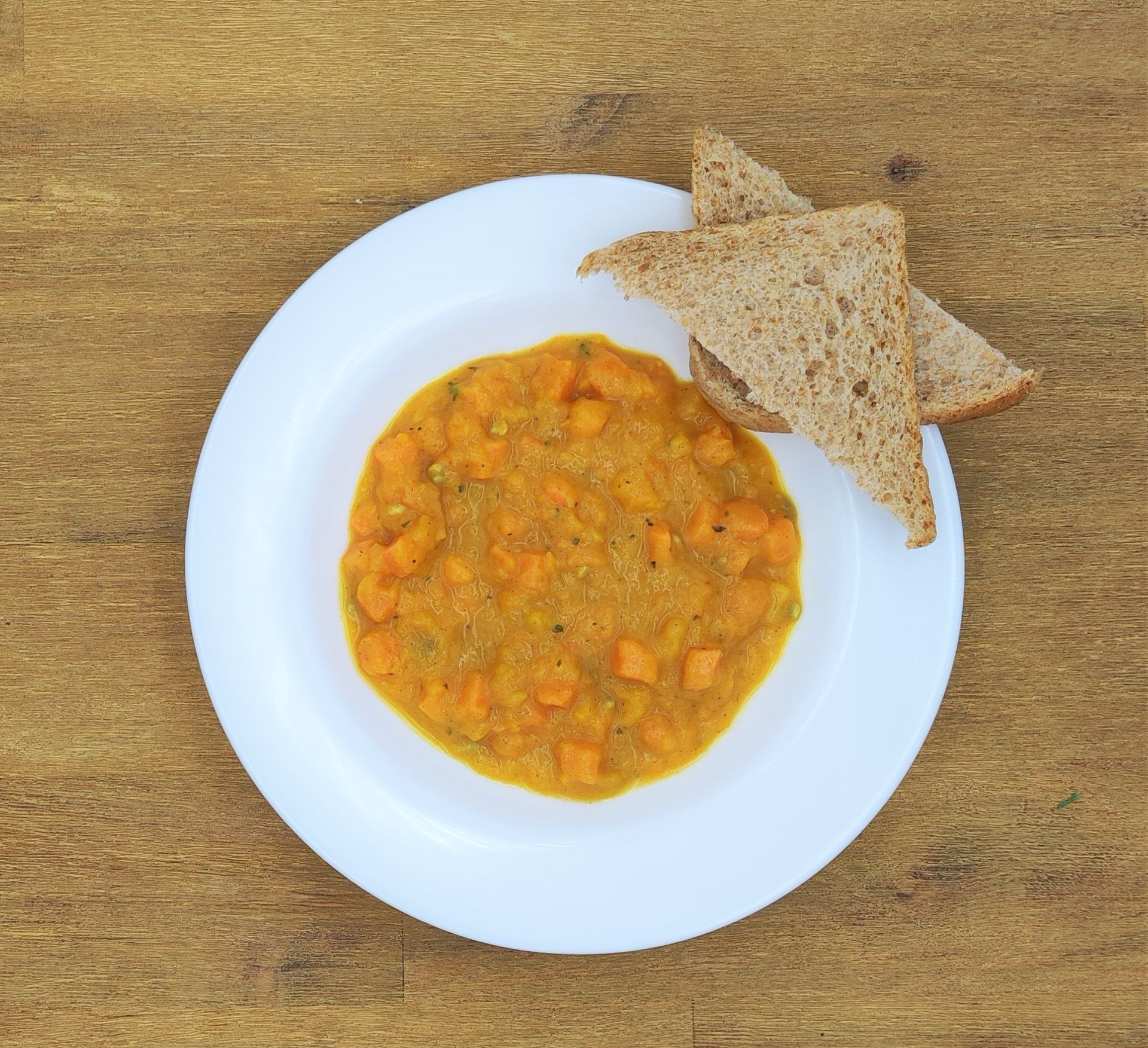 Suppe mit gewürfeltem Gemüse auf einem weißen Teller mit zwei Scheiben Toast auf einer Holzplatte.