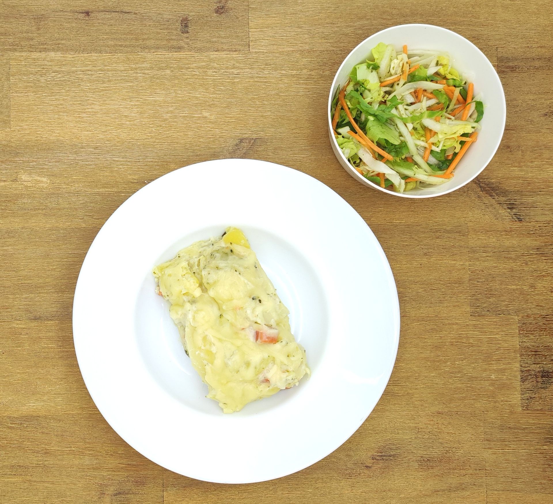 Teller mit abgedecktem Essen und Schüssel mit Salat auf einer Holzoberfläche.